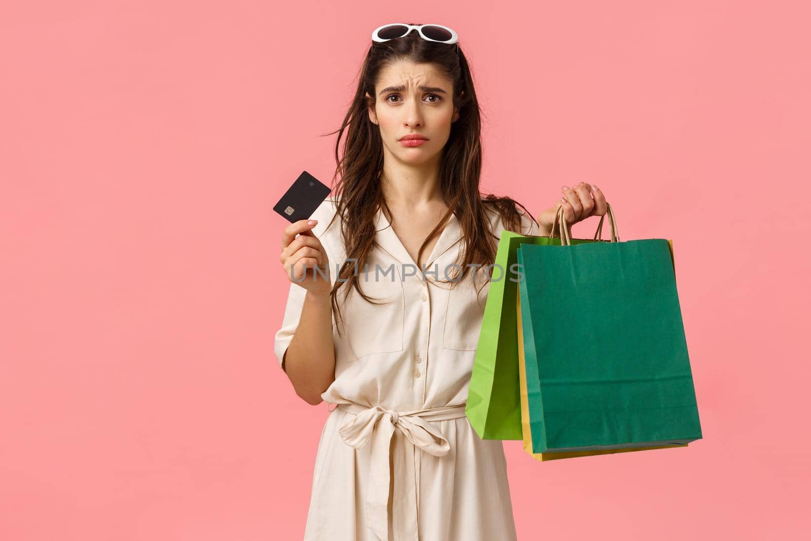 Upset and distressed young brunette female feeling sad spent all money, looking uneasy and concerned at credit card, holding shopping bags, standing pink background gloomy by Benzoix