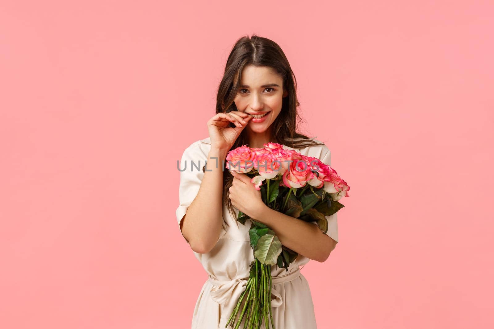 Romantic date, beauty and fashion concept. Gorgeous brunette girl in dress, receive beautiful bouquet flowers, got roses, biting finger and gazing camera flirty, standing pink background.