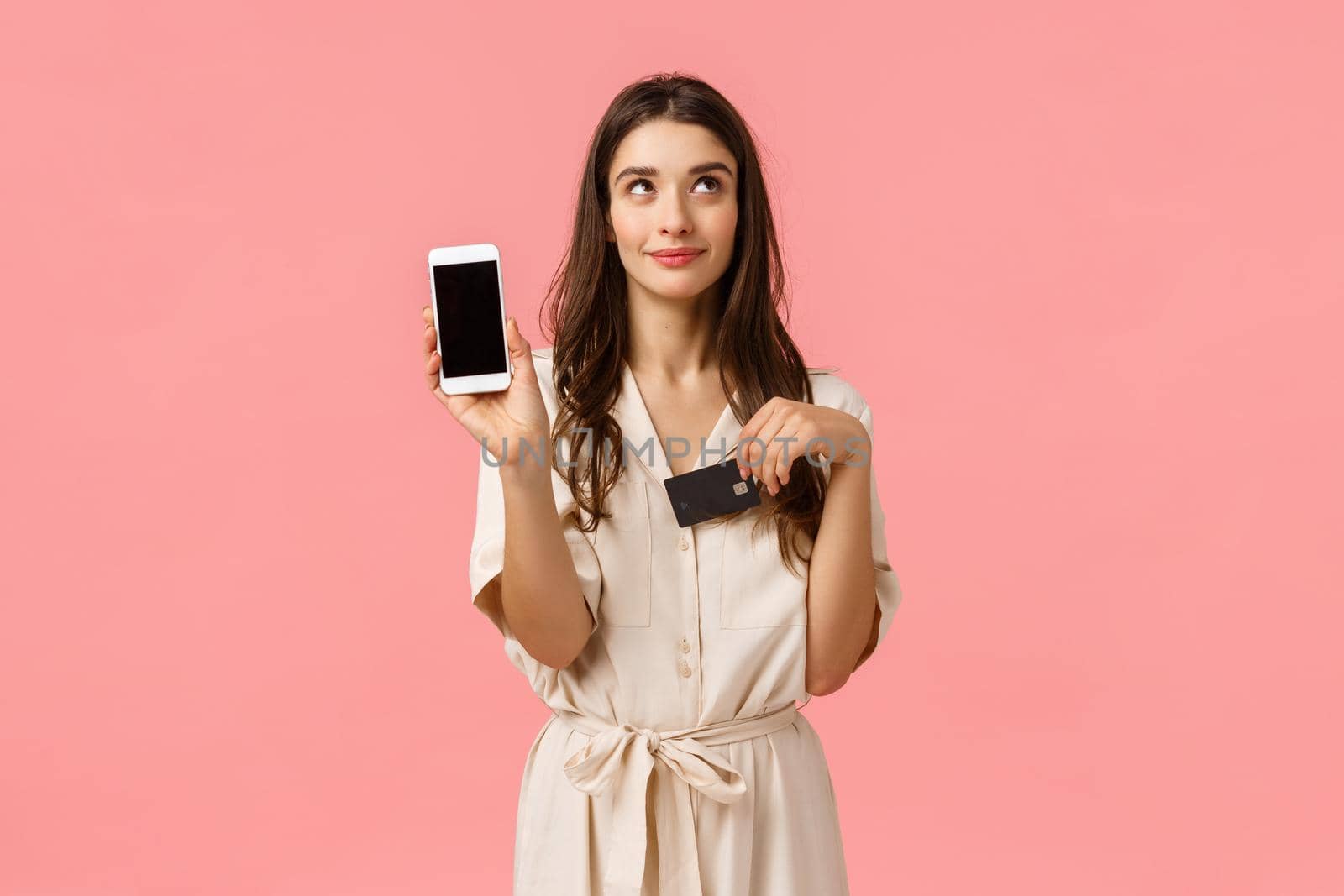 Dreamy and coquettish good-looking young woman in dress, dreaming waiting for delivery order online, holding smartphone and credit card, showing mobile screen, standing pink background.