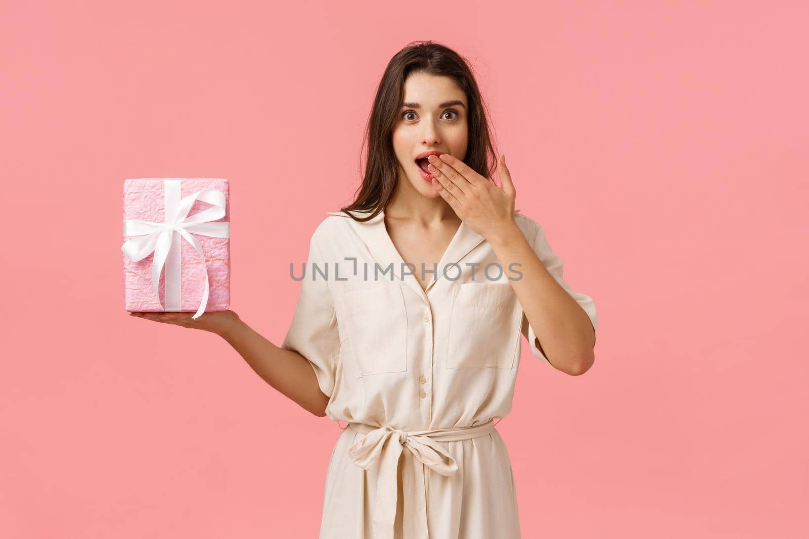 Surprises, holidays and celebration concept. Amused and cheerful attractive young brunette girl in dress, gasping open mouth amazed, receive wonderful birthday gift, pink background.