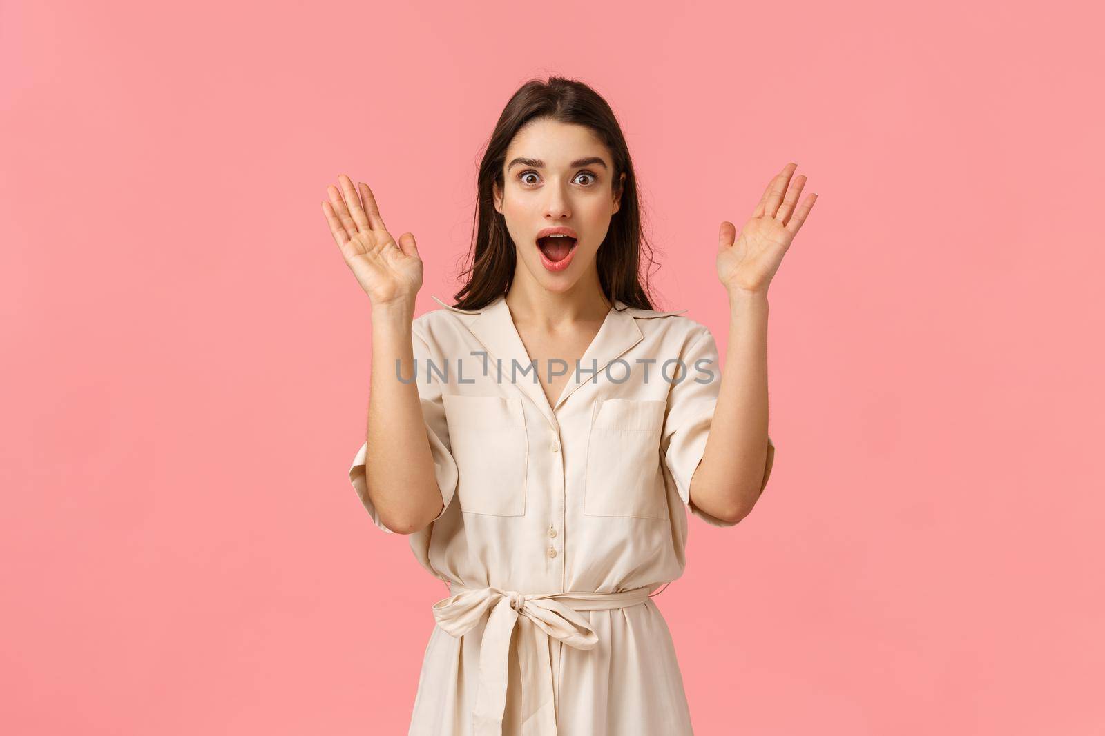 Wow super surprise. Amazed and wondered, excited happy, glad pretty caucasian woman in cute dress, raise hands up astonished, open mouth and looking camera thrilled, standing pink background.