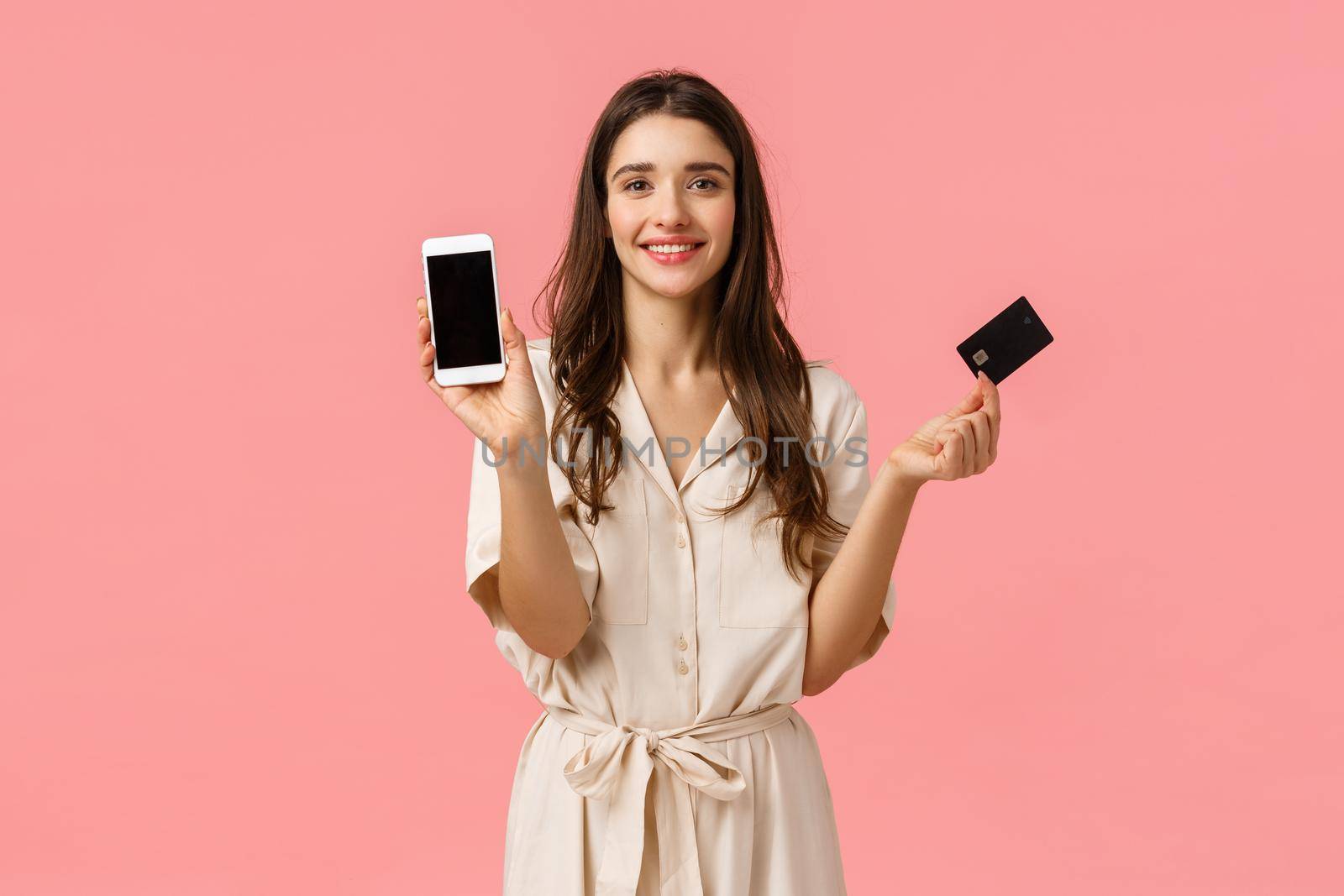 Presents, shopping and beauty concept. Fashionable young woman in trendy dress, holding smartphone and credit card, smiling cheerfully, order product online, standing pink background.