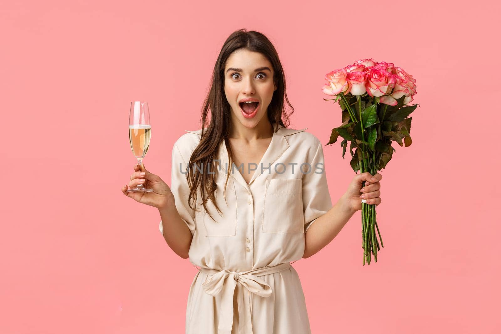 Wonderful news, celebration and emotions concept. Cheerful attractive brunette female in dress, spread hands sideways surprised and wondered, open mouth gasping, hold champagne glass and flowers by Benzoix
