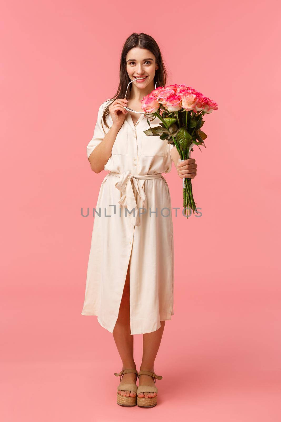 Full-length vertical portrait sensual, romantic and coquettish young woman seeing something interesting, looking with temptation or desire, holding bouquet flowers, received roses, pink background by Benzoix