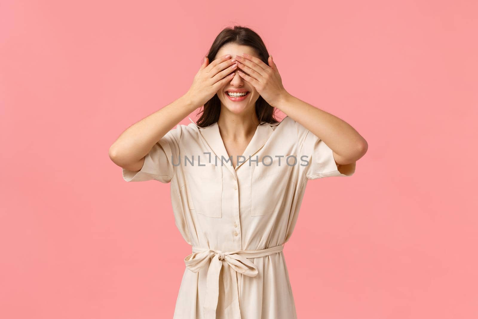 Romantic moment, beauty and tenderness concept. Attractive brunette girlfriend asking close eyes, cover sight with palms as waiting surprise gift, standing pink background, smiling with temptation by Benzoix