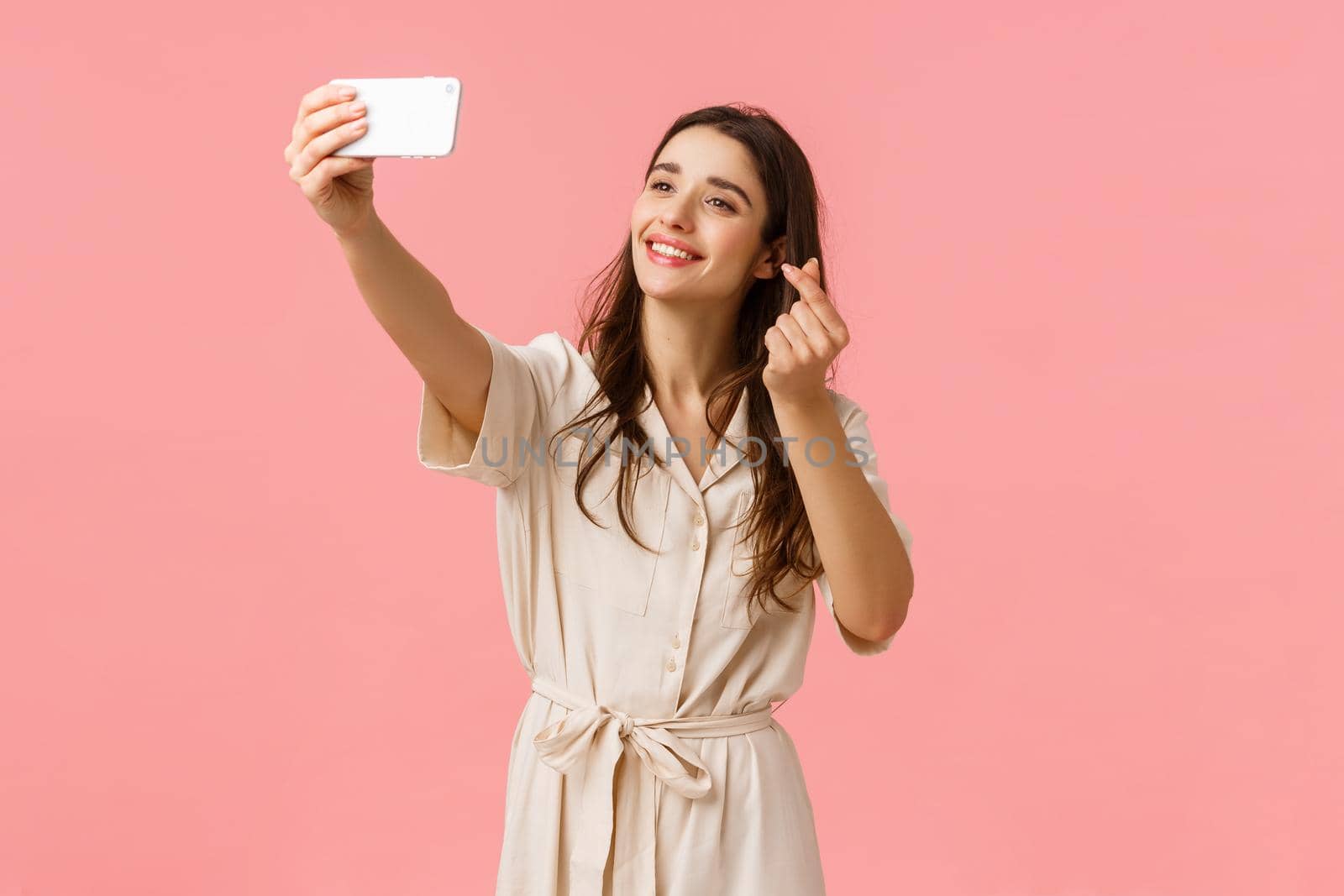 Cheerful lovely young european woman using filter to take cute selfie, photographing on mobile camera, smiling and showing korean heart gesture at smartphone, standing pink background.