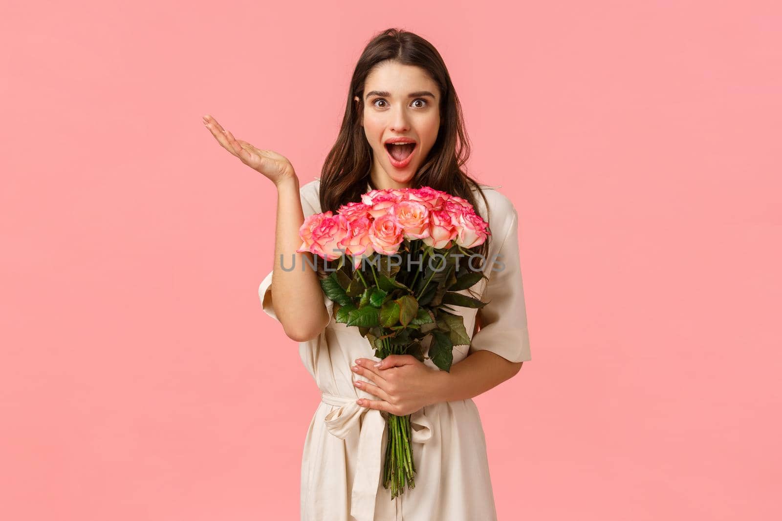 Wow such wonferful unexpected surprise. Charming lovely and romantic cute brunette girl in dress over pink wall, receive beautiful flowers, holding roses and looking amused, raise hand astonished by Benzoix