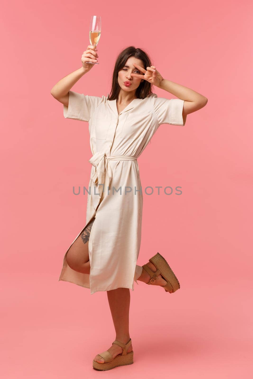 Full-length vertical portrait carefree, happy gorgeous woman dancing, enjoying the night, having party raising champagne glass and show peace sign over eye, fold lips in kiss, pink background.
