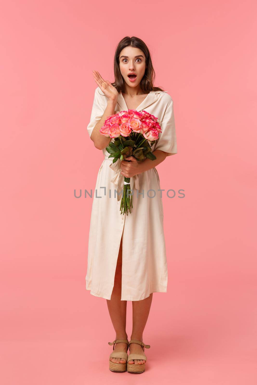 Full-length vertical portrait pretty surprised girl receive unexpected gift, got delivery holding flowers, gasping open mouth amazed and looking speechless with stunned expression, pink background.