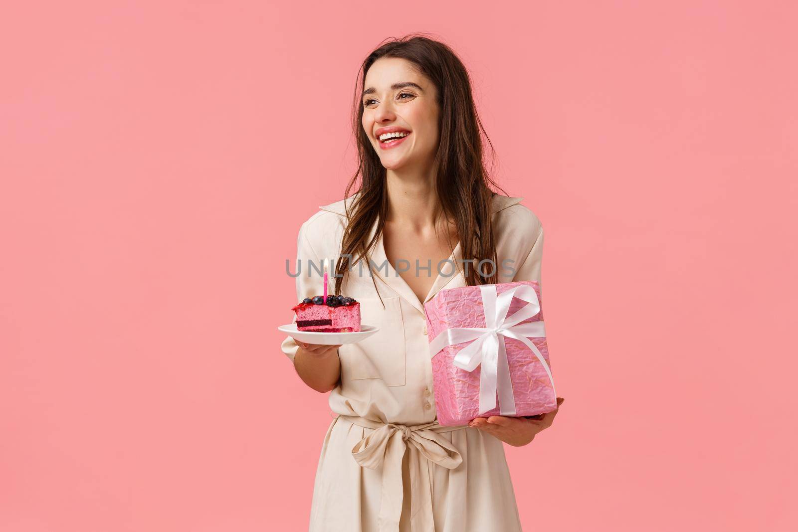 Celebration, party and happiness concept. Carefree smiling happy european woman having fun parying own b-day party, holding gift and piece cake, blow-out candle, pink background by Benzoix