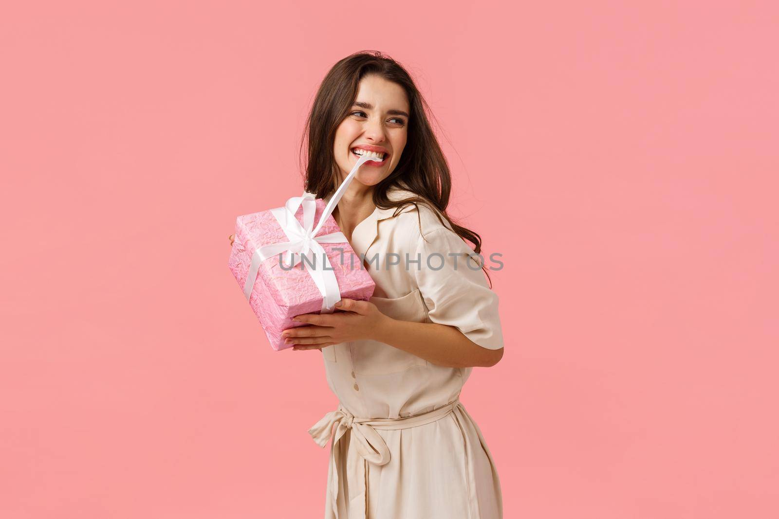 Happiness, tenderness and celebration concept. Carefree happy and upbeat young girl partying, having amazing birthday day, biting knot on cute gift, smiling, tempting unwrap present, pink background.