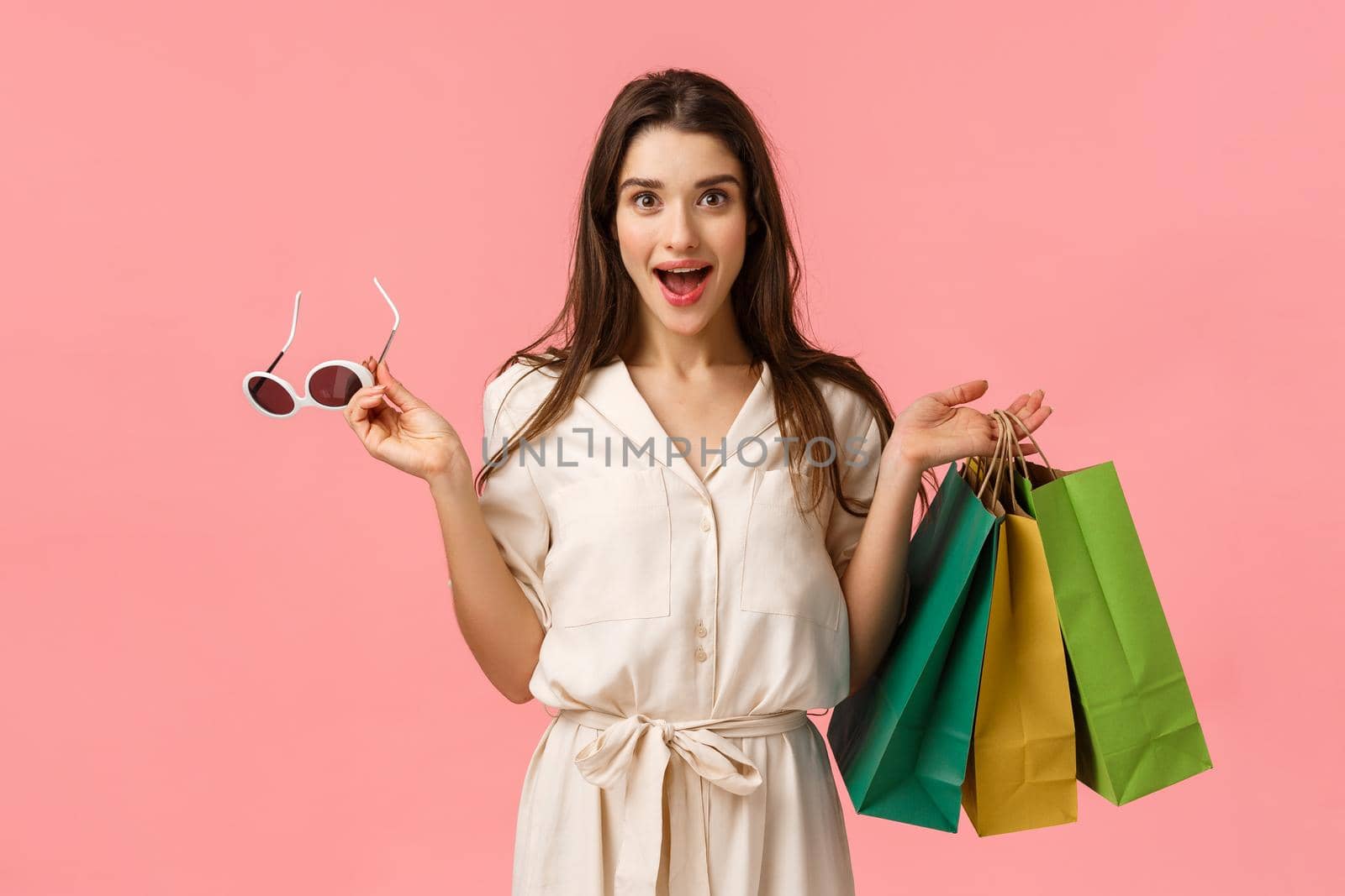 Cheerful gorgeous caucasian woman in dress, holding shopping bags and sunglasses, smiling intrigued and excited, seeing beautiful garment on stalls want buy, standing pink background by Benzoix