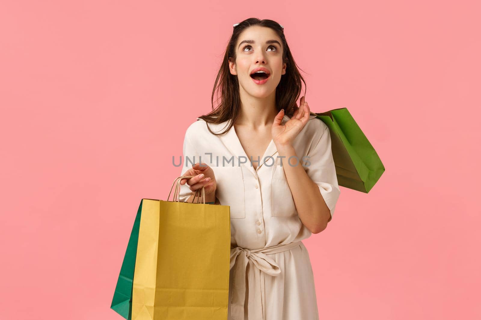 Dreamy excited and amused cheerful brunette girl in dress, holding shopping bags, looking up astonished and amazed, smiling wondered, feeling happy strolling in city malls. Copy space