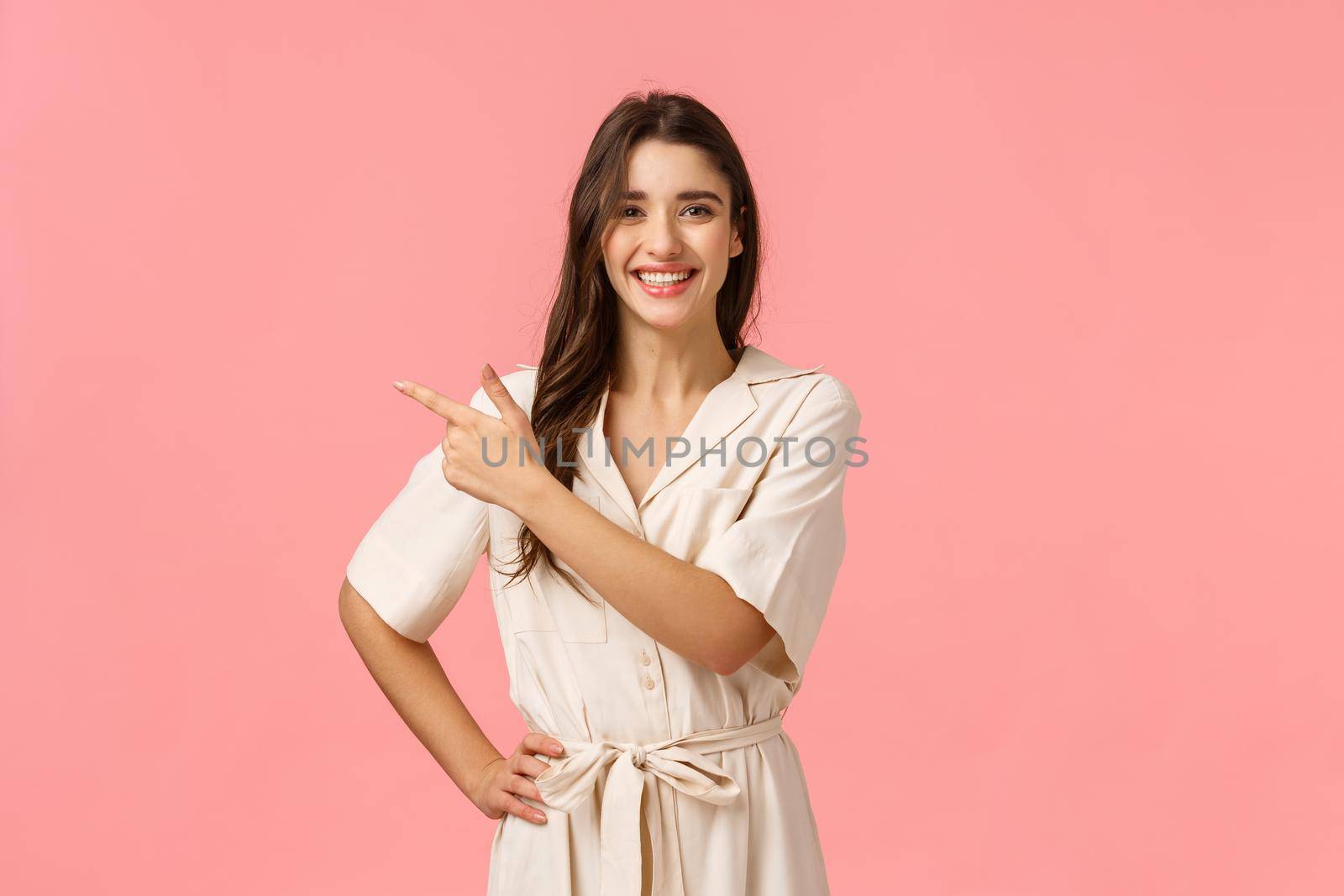 Enthusiastic positive cute brunette girl in pretty dress, pointing upper left corner, gladly showing product discounts promo, standing pink background, smiling joyfuly and excited.
