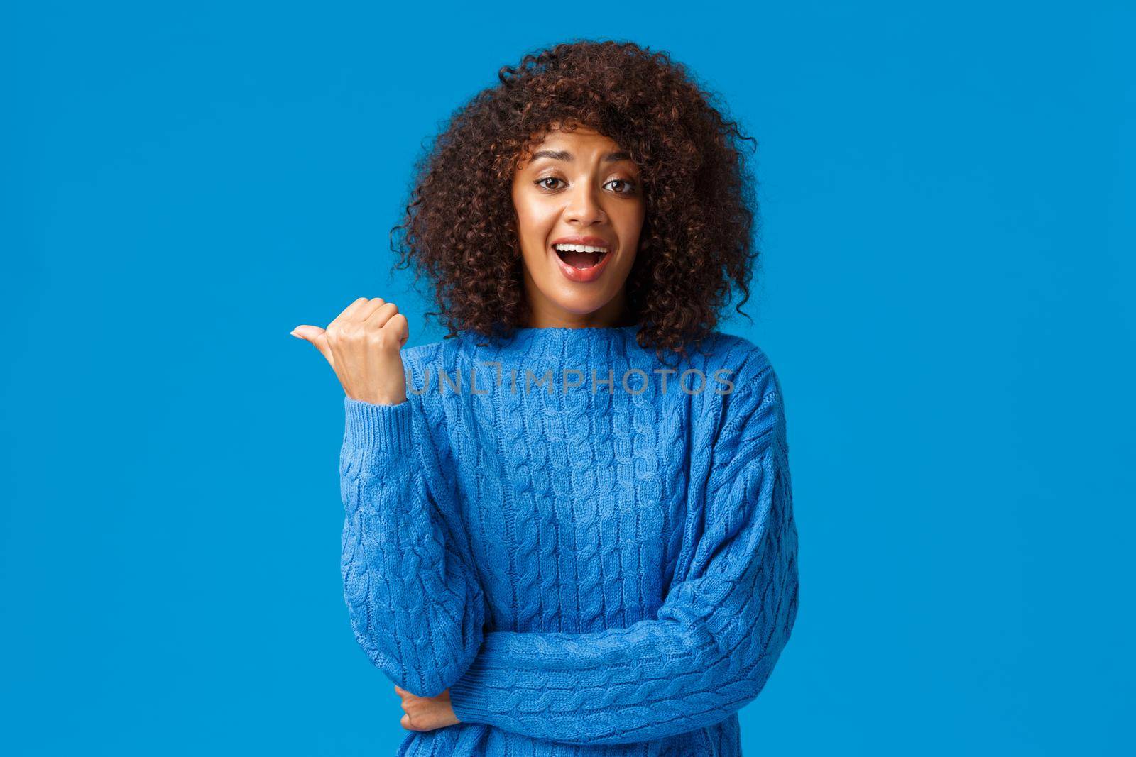 Excited happy pretty african-american woman with afro haircut in winter sweater, pointing thumb left and laughing as discuss funny moment, talking casually during party, blue background.