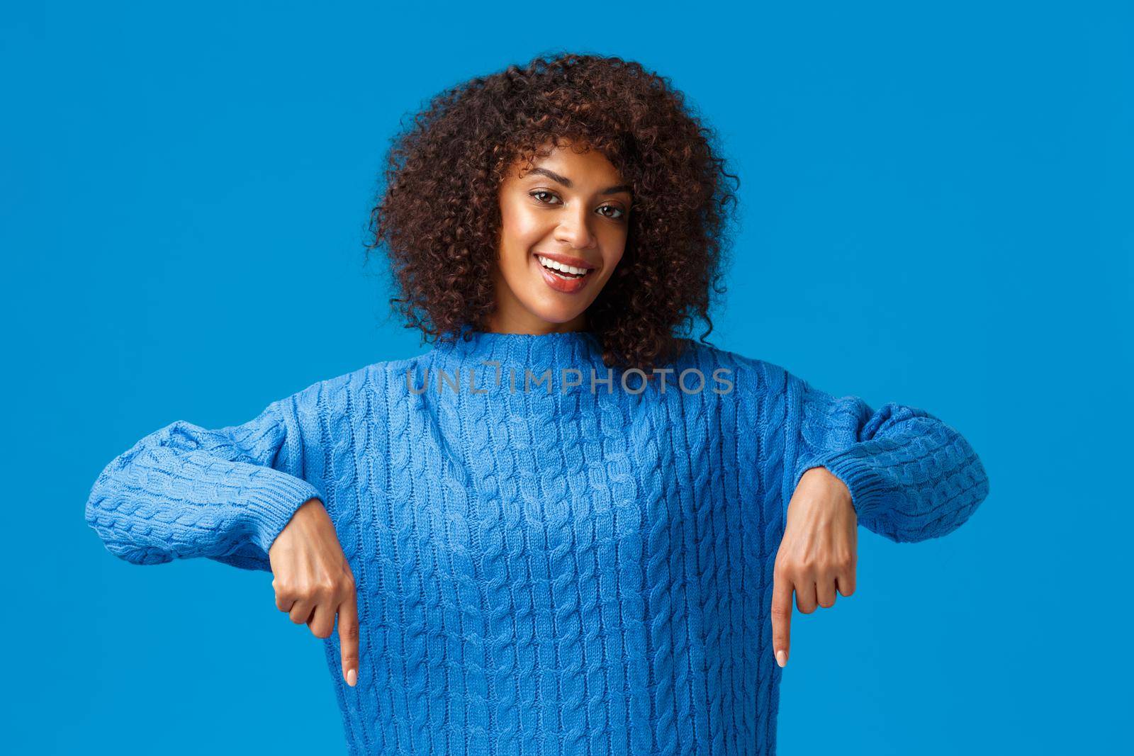 Lovely, friendly sensual young african-american woman in winter sweater, pointing down, inviting brows through awesome website shopping online, indicating bottom advertisement, blue background.