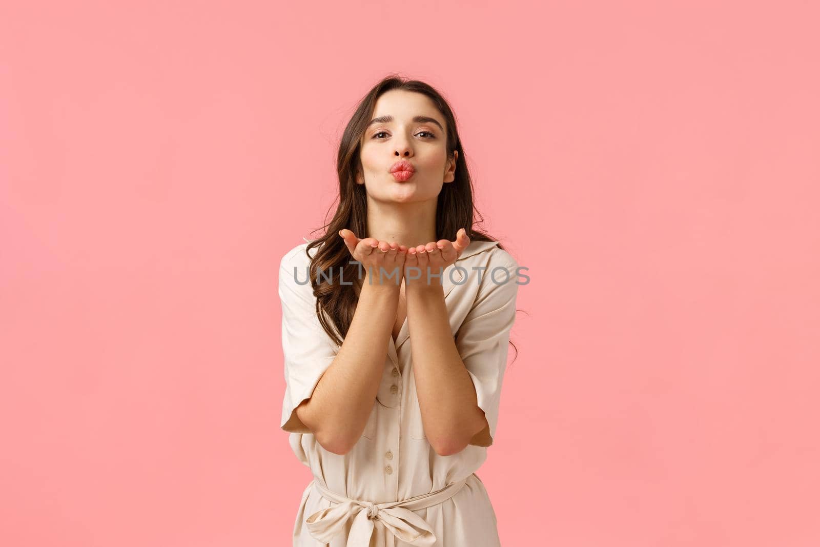 Sending love and care. Charming cute and tender european brunette with curly hair, wearing spring dress, leaning with hands near folded lips, blowing air kiss with love, standing pink background by Benzoix