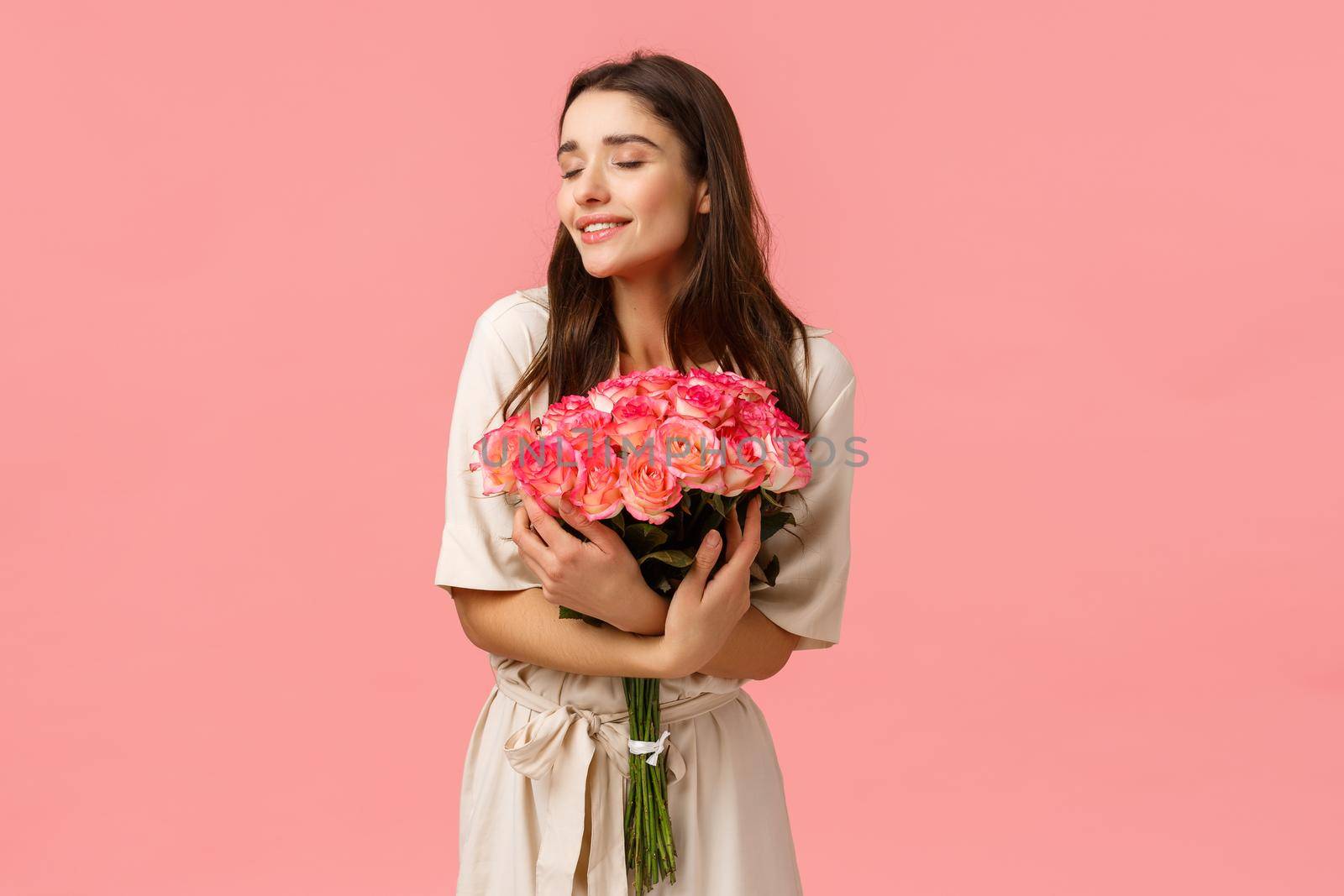 Tenderness, beauty and romance concept. Romantic good-looking tender brunette girl in dress, smiling sensually and delighted, holding roses, receive bouquet roses and laughing, pink background by Benzoix