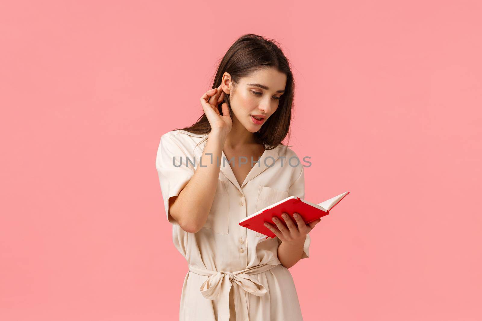 Romantic and tender young alluring woman in dress, put hair strand behind ear and reading notes from red notebook, writing something, studying for exams, standing pink background. Copy space