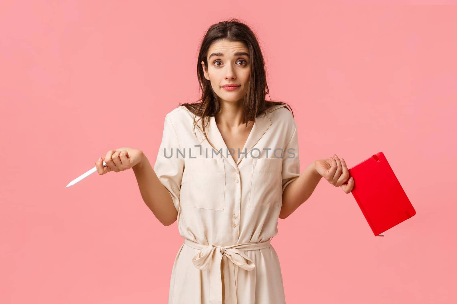 Confused and indecisive beautiful young brunette female in dress, dont know anything, shrugging and spread hands sideways making clueless expression, holding notebook and pen, pink background.