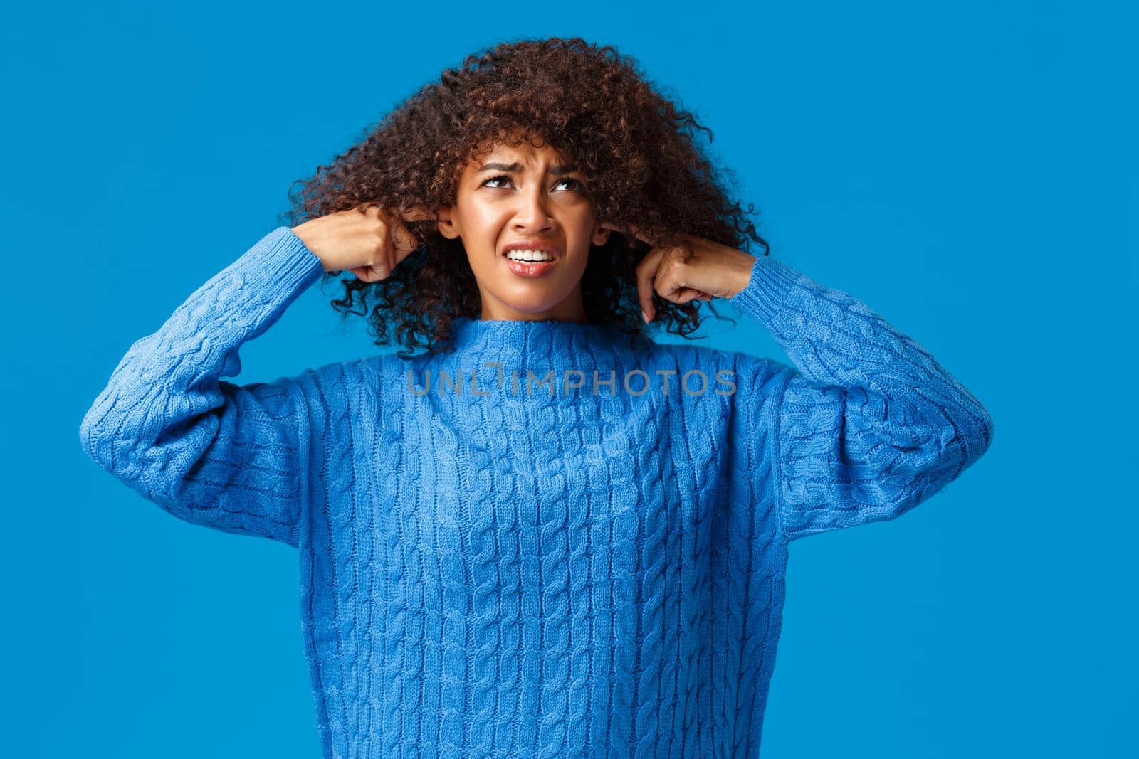 Bothered and interrupted displeased african-american college student female in winter sweater, looking up and grimacing, shut ears with fingers, not hear loud awful music, neigbours having party by Benzoix