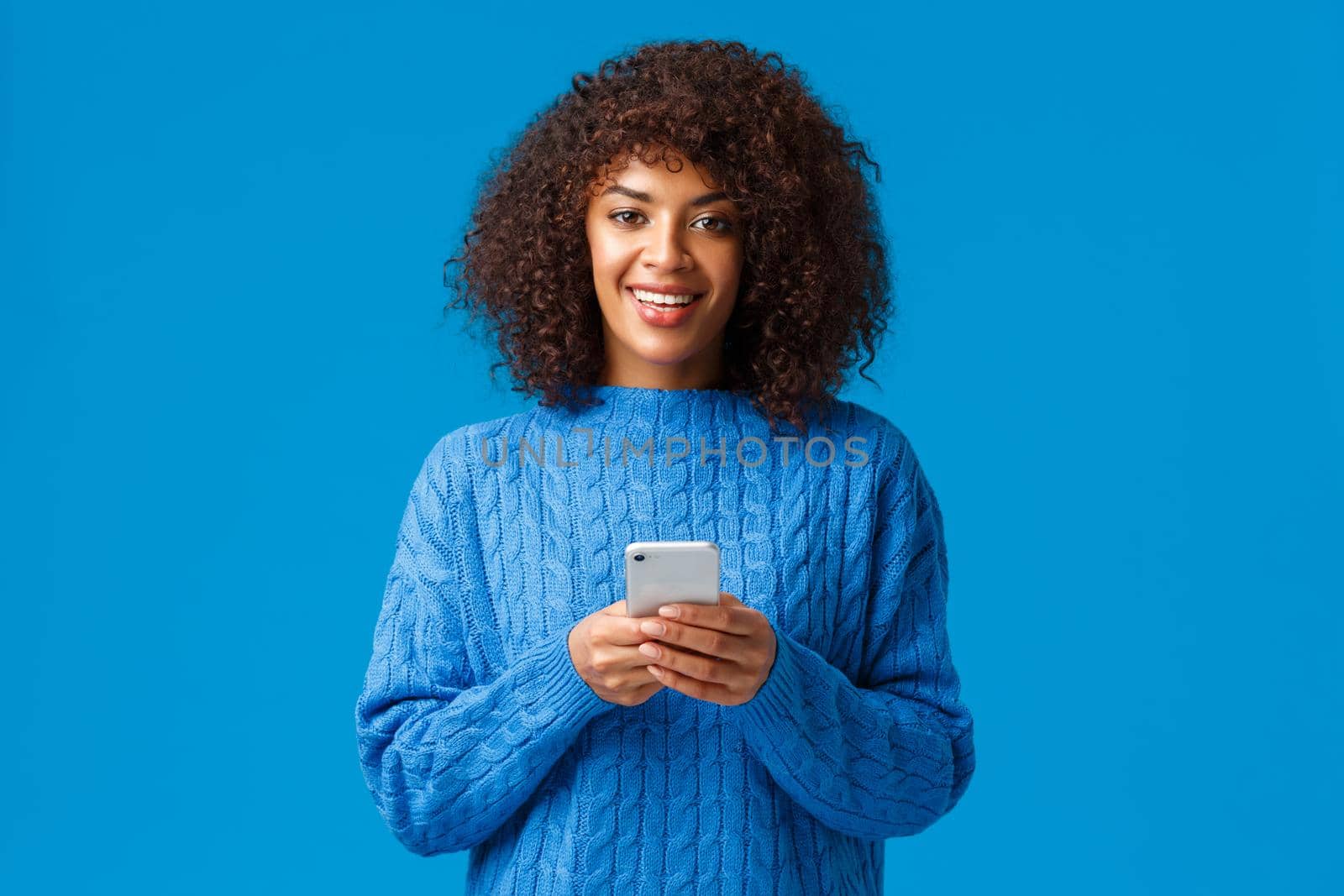 Smartphone addication, gen-z and people concept. Charismatic lovely smiling african-american woman with afro haircut in winter sweater, holding smartphone and looking camera, blue background.