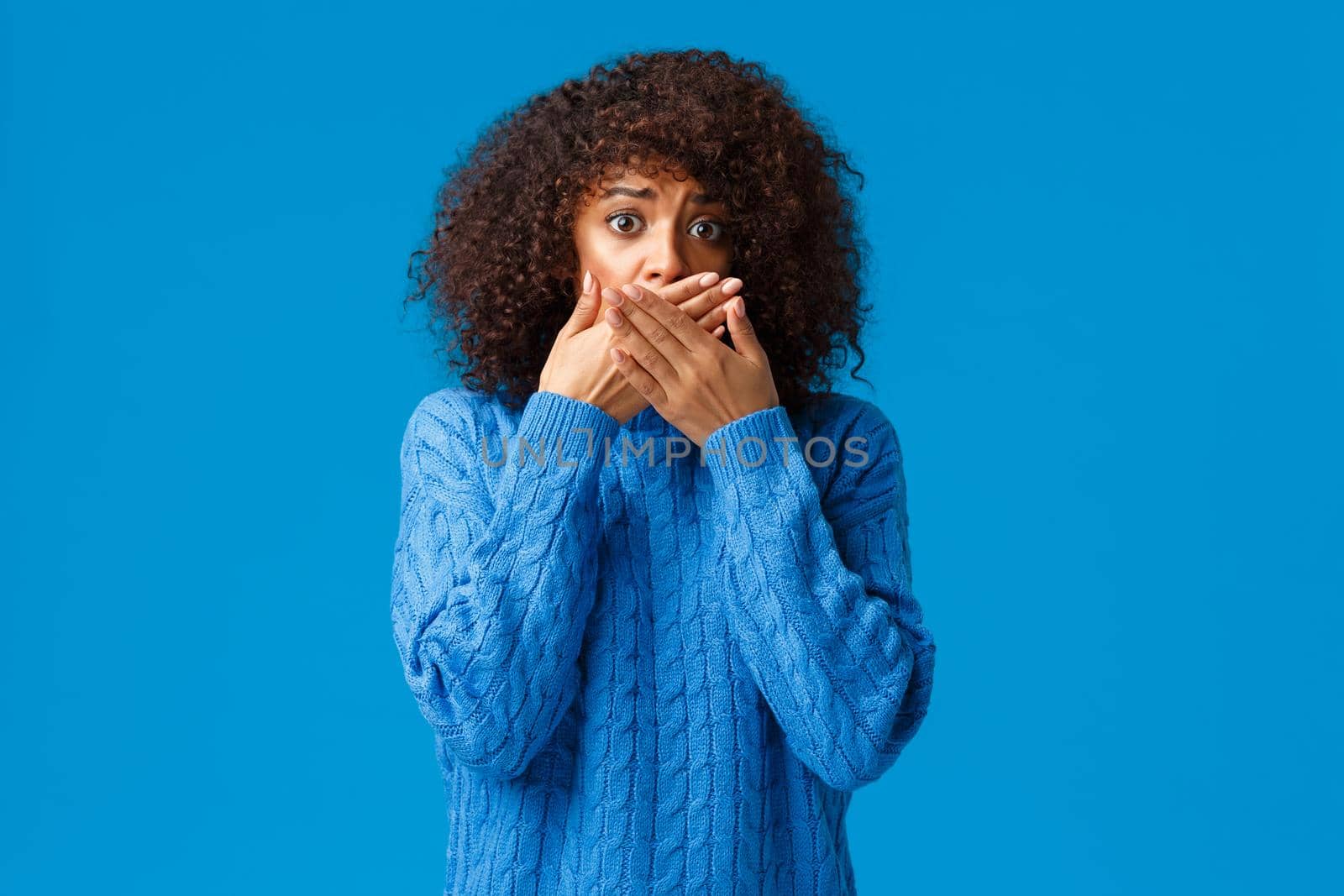 Scared and timid, insecure anxious young african-american woman witness terrible scene, gasping hold hands over mouth not scream, staring in panic and nervously standing over blue background.