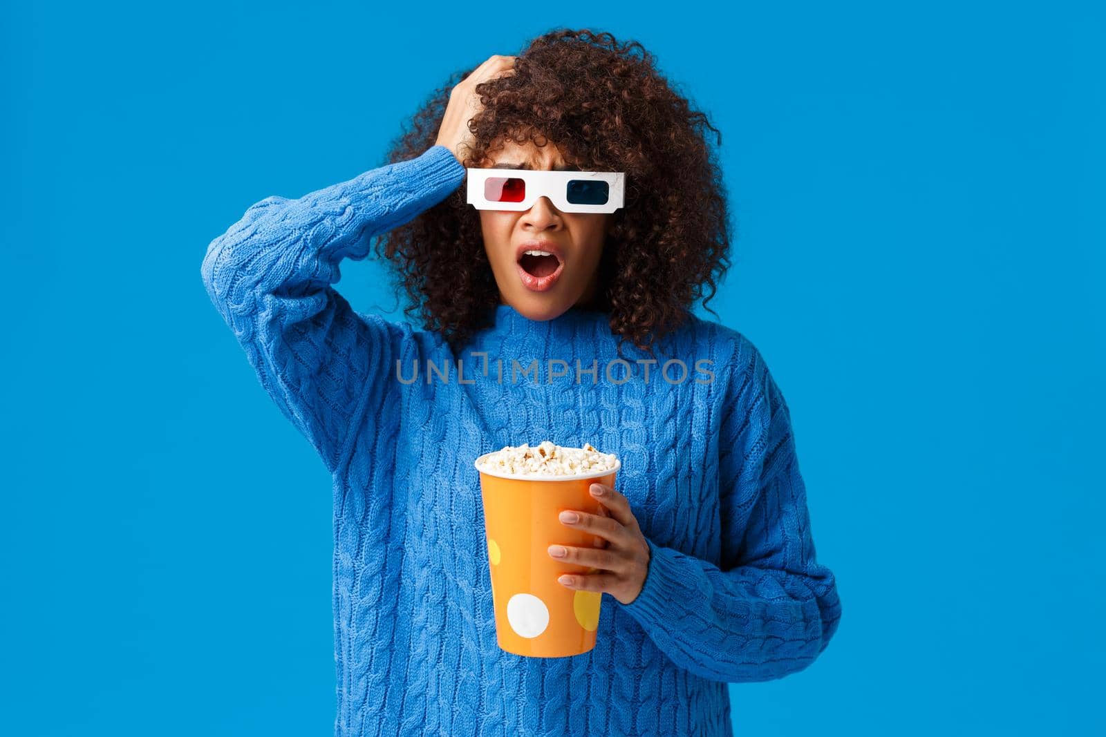 Shocked african-american pretty woman concerned with shocking cliffhanger in movie, grab head uneasy and upset, drop jaw, holding popcorn watching film in cinema, blue background by Benzoix