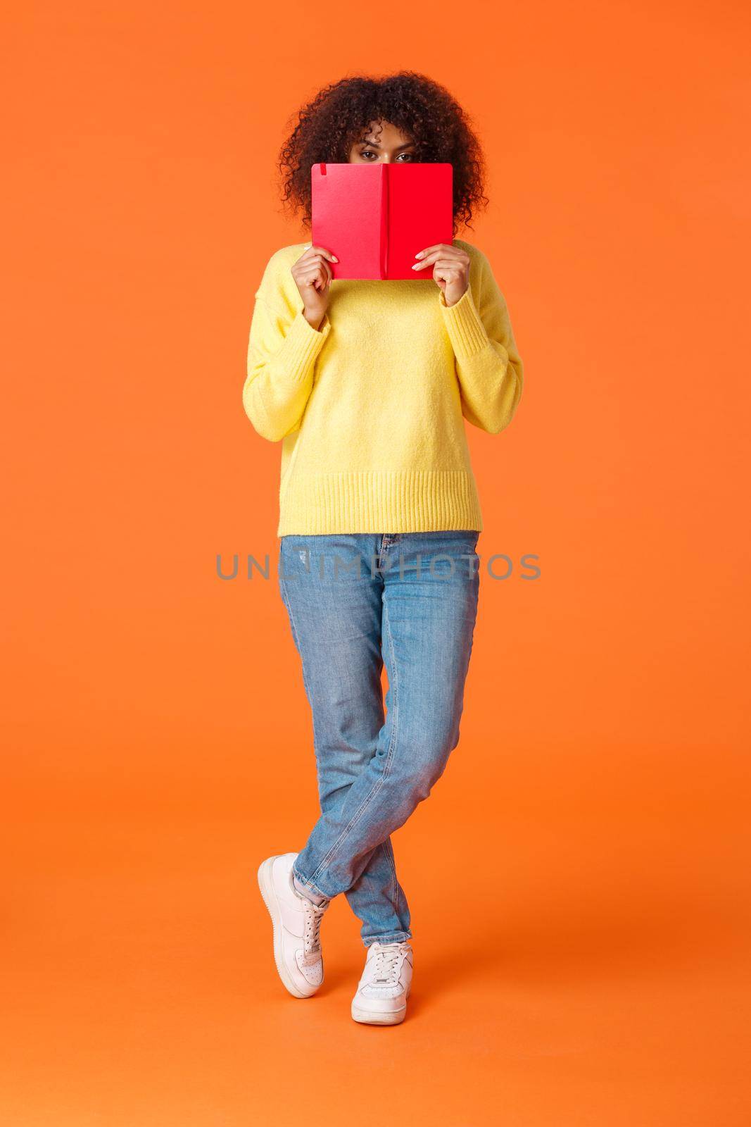 Full-length vertical shot shy and cute african-american modern urban girl with afro haircut, hiding face behind red notebook and peeking camera, writing in diary, standing orange background by Benzoix