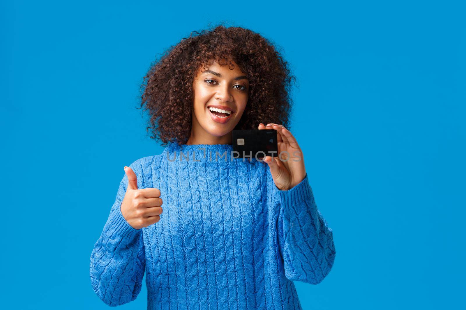 Happy, satisfied customer recommend bank. Attractive modern young african american woman in winter sweater buying everything with credit card, show thumb-up gesture, blue background.