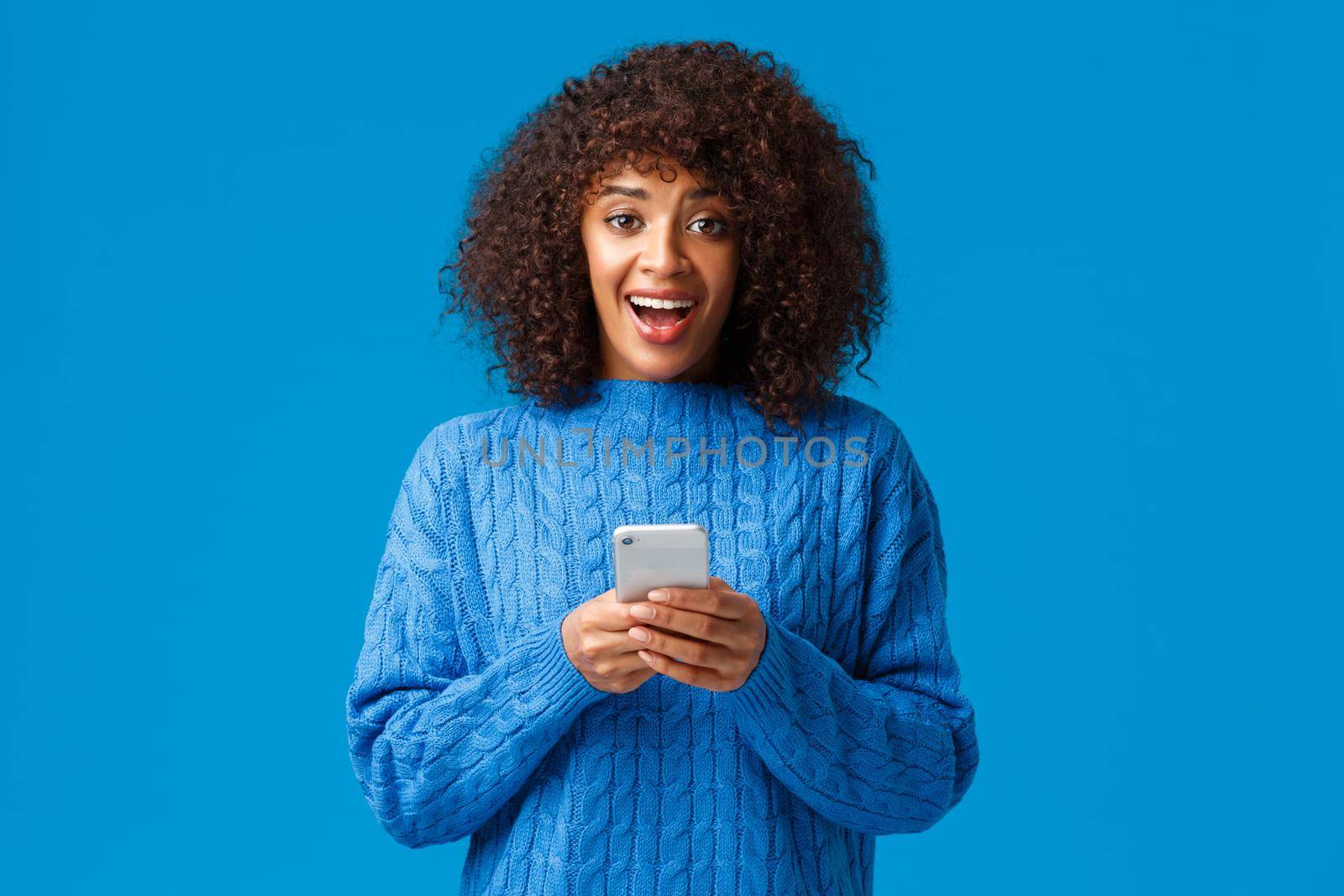 Excited and overwhelmed smiling attractive young african-american woman, wearing winter blue sweater, looking amazed and surprised camera as receive great news over smartphone message.