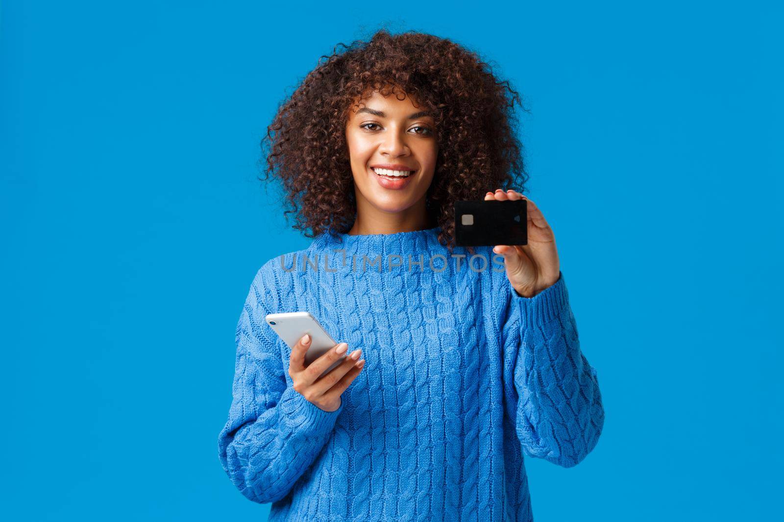 Cute pleased african-american female bank customer recommend credit card and banking system service, holding smartphone and smiling, buying online, shopping in internet stores, blue background.