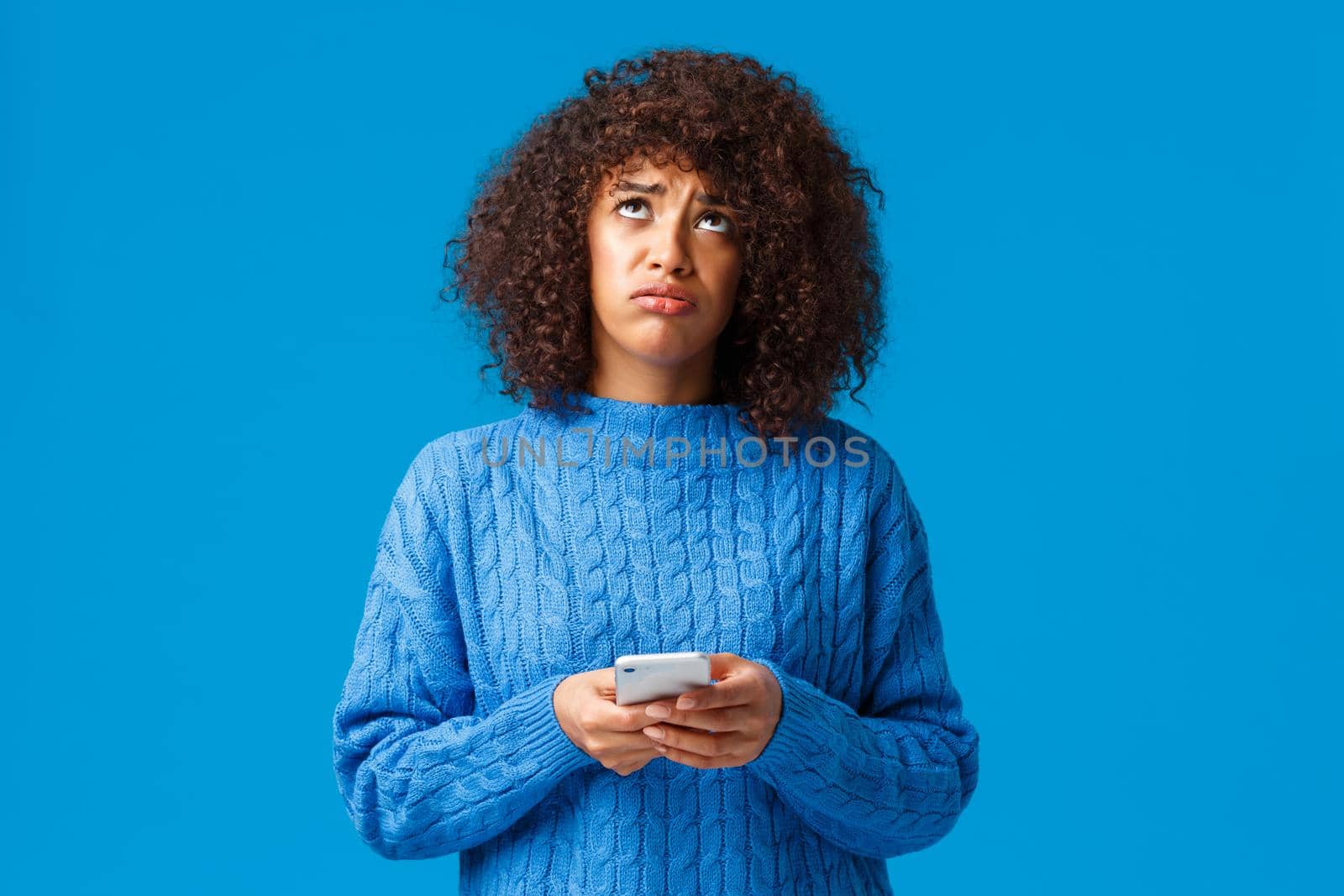 Upset and uneasy african-american gloomy girl feeling like loser, looking up at sky asking god why, sighing sad, holding smartphone, standing troubled and sulking over blue background.