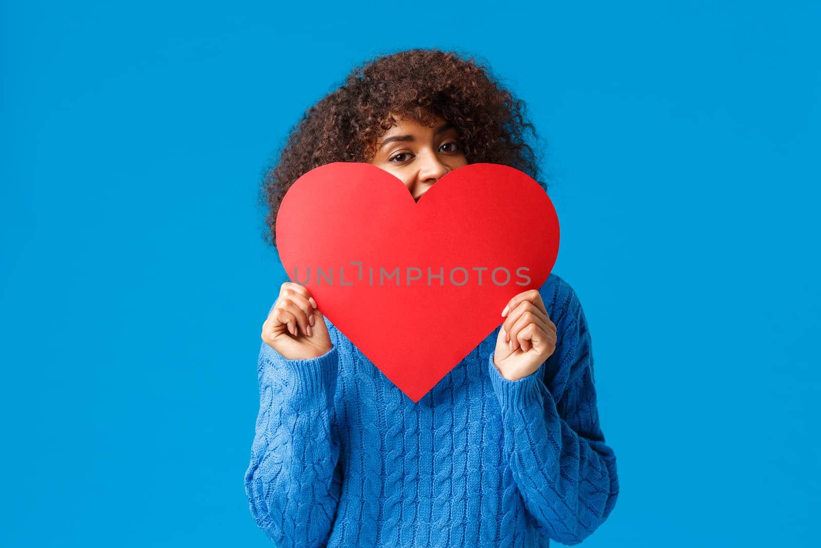 Cute and lovely flushed african-american girl, with afro haircut, in sweater, hiding face behind big red heart and peeking joyfully camera, confessing in love, sympathy, standing blue background.