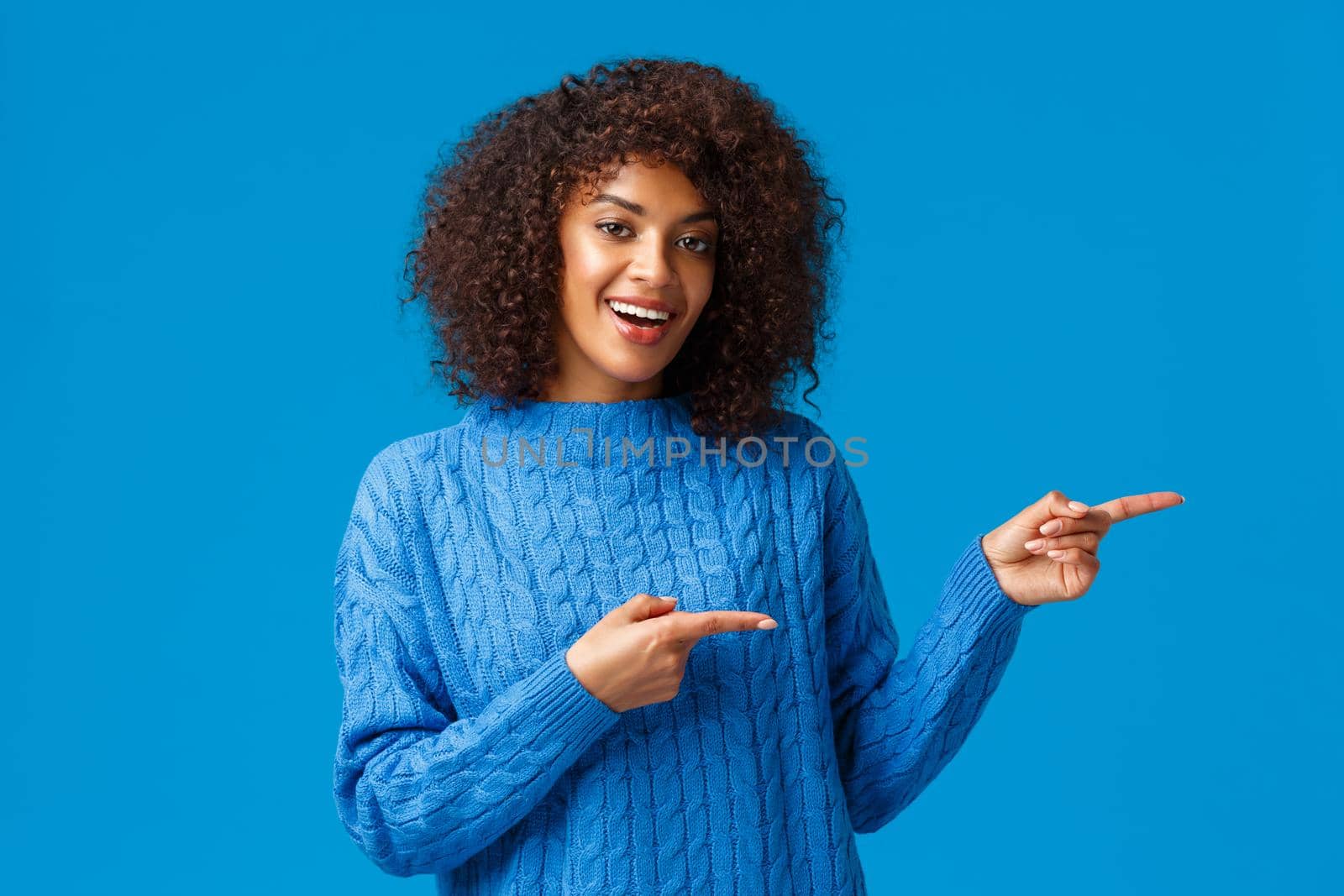 Hey visit this site. Cheerful happy african-american female in winter sweater, inviting check-out holiday prices and special discounts, pointing fingers right side and smiling, blue background.