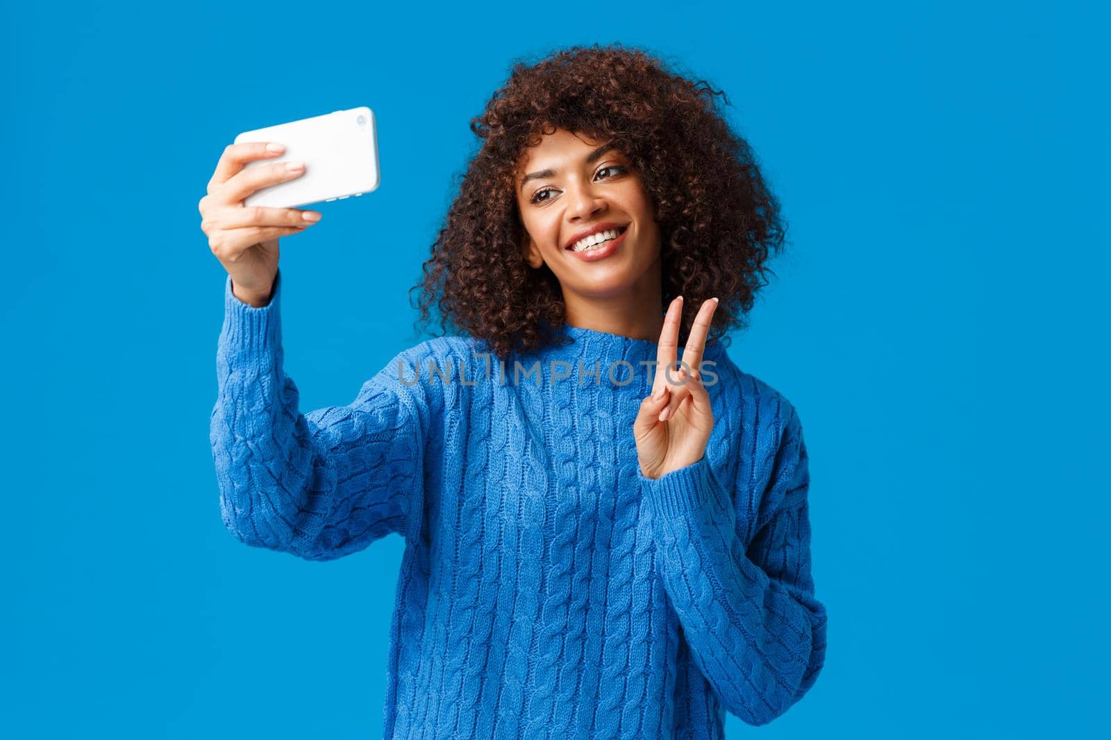 Cheerful friendly and cute african-american female student taking photo herself apply filters in new smartphone app, taking selfie tilt head lovely smiling, making peace gesture, blue background by Benzoix