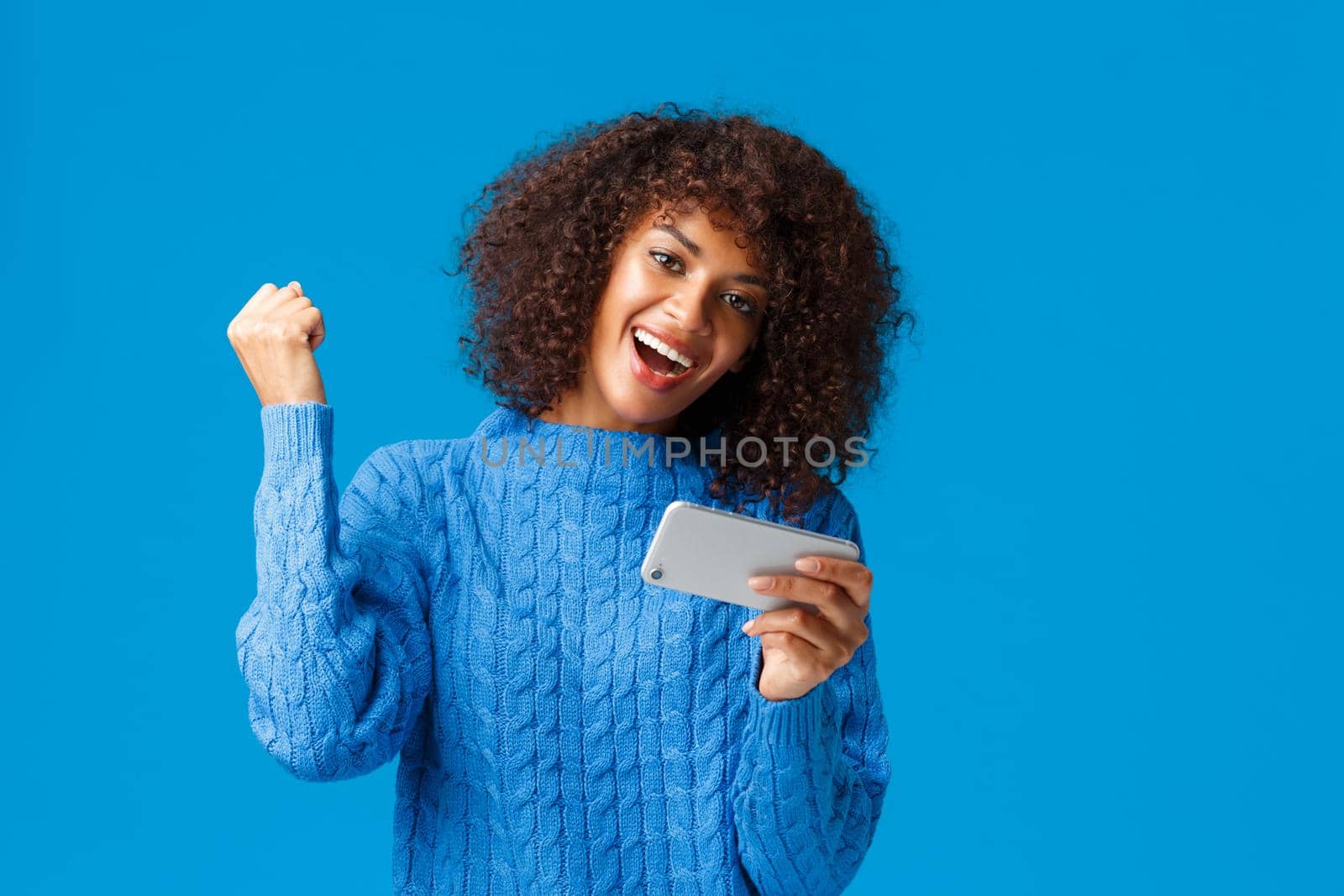 Excited happy lucky african-american woman passed level, finished race first in game, holding smartphone playing app, cheering, fist up like champion, celebrating victory, blue background.