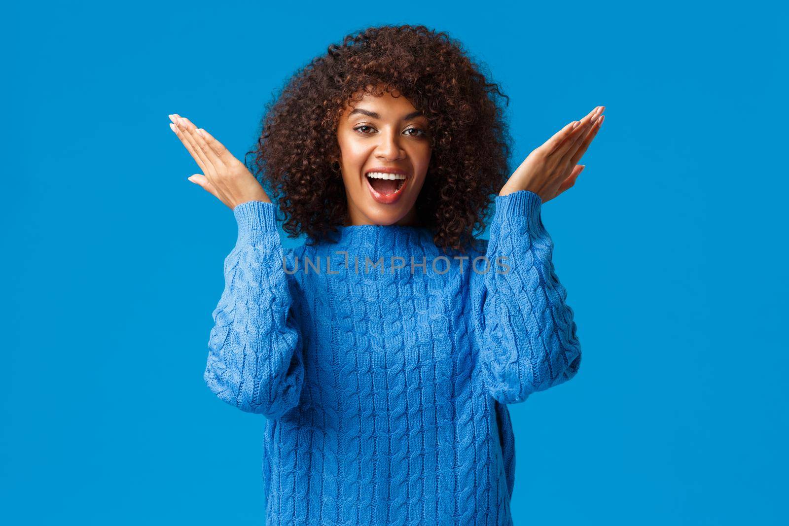 Cute and playful, funny happy african-american woman with afro haircut in winter sweater, open eyes to see holiday surprise, valentines day secret gift, staring amused and delighted, blue background.