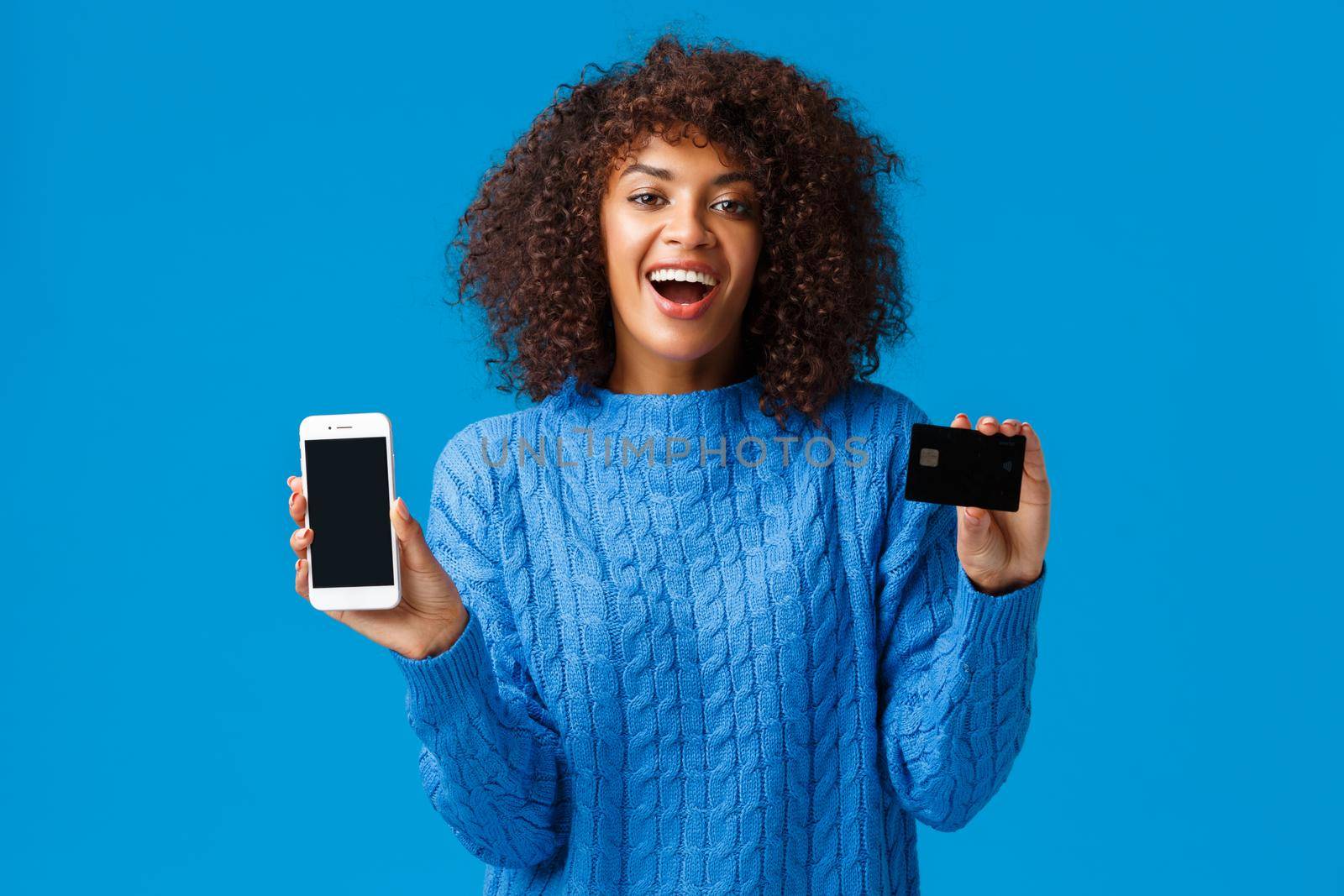 Happy and excited, charming african-american female shopaholic buying presents online, showing application, shopping app and credit card, smiling and laughing, standing blue background.