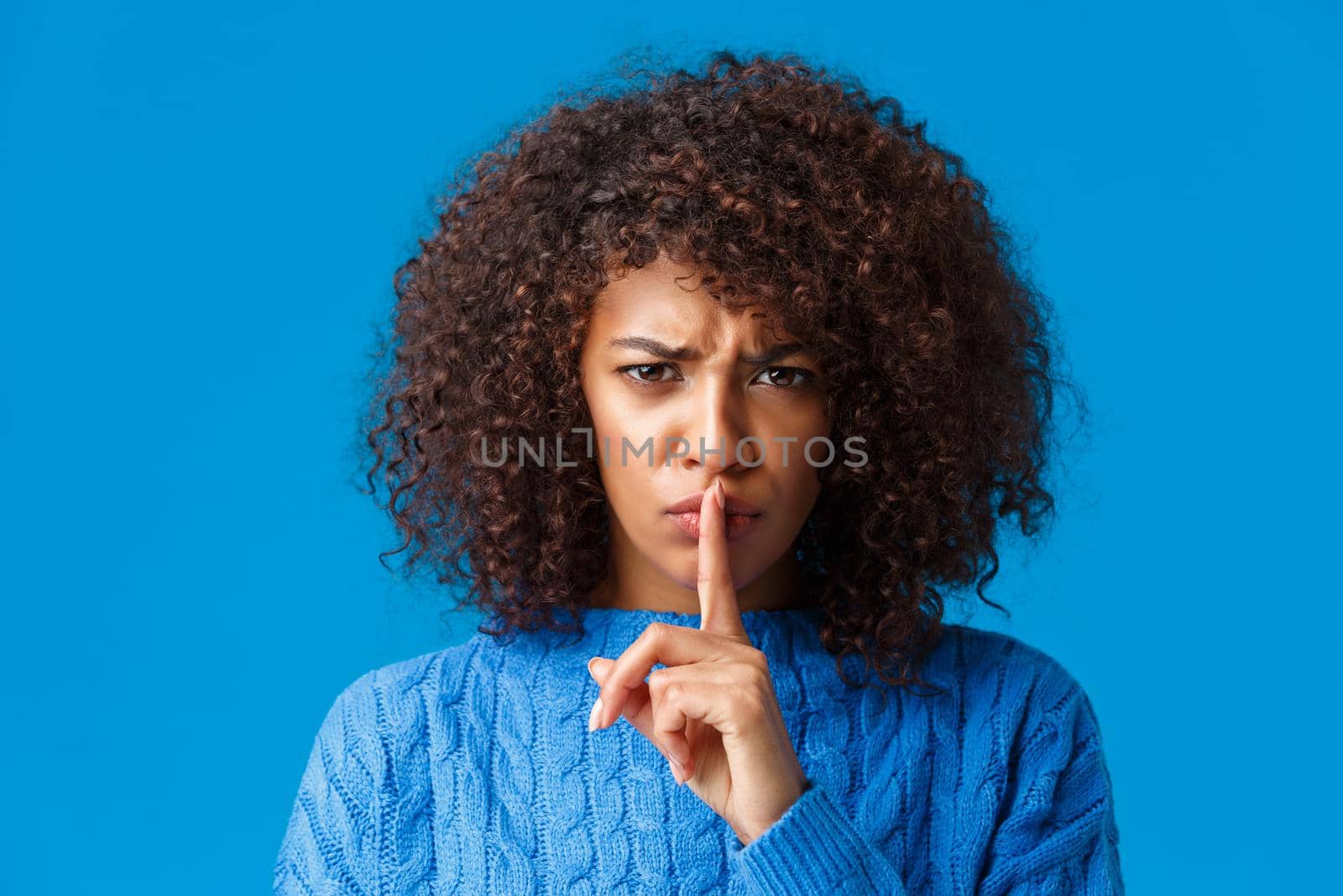 Close-up portrait offended and upset silly timid african-american girl frowning displeased, shushing angry at camera with index finger pressed lip, demand silence, asking keep quiet, blue background.