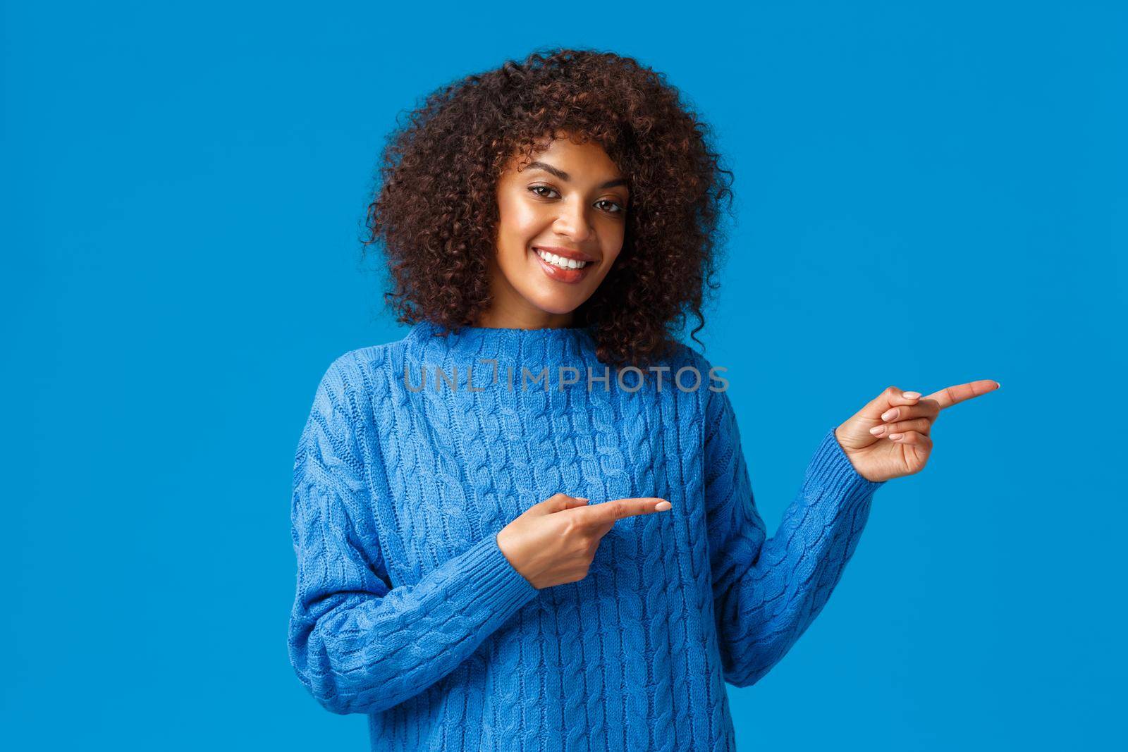Lovely adult african-american female with afro haircut, pointing fingers right, suggesting go direction, visit site or download app, presenting great product and smiling satisfied, blue background.