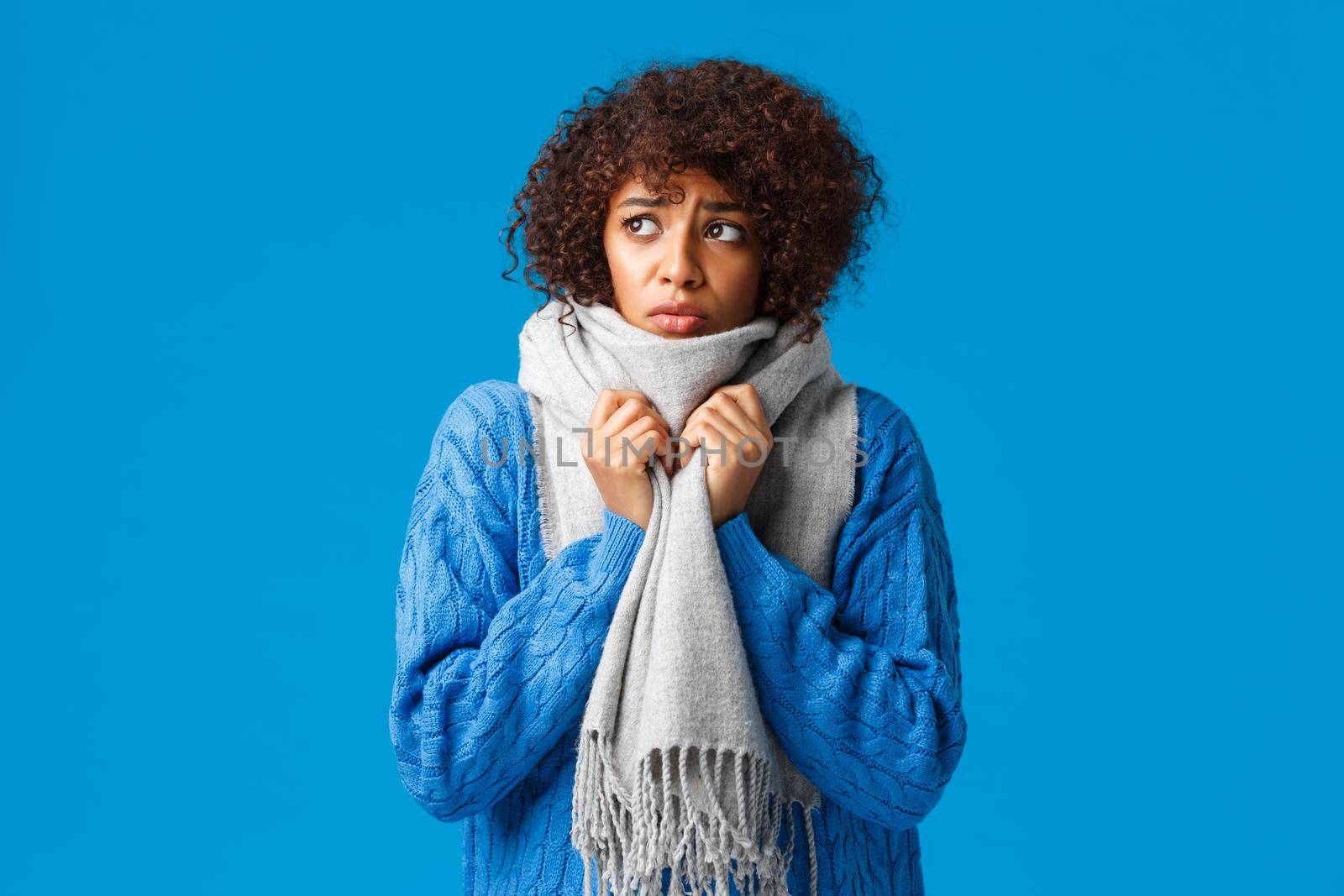 Silly and sad, gloomy african-american girl looking outside windy snowy weather winter, wrap neck with warm scarf, tremble from cold feeling discomfort from low tempreture at home, blue background.
