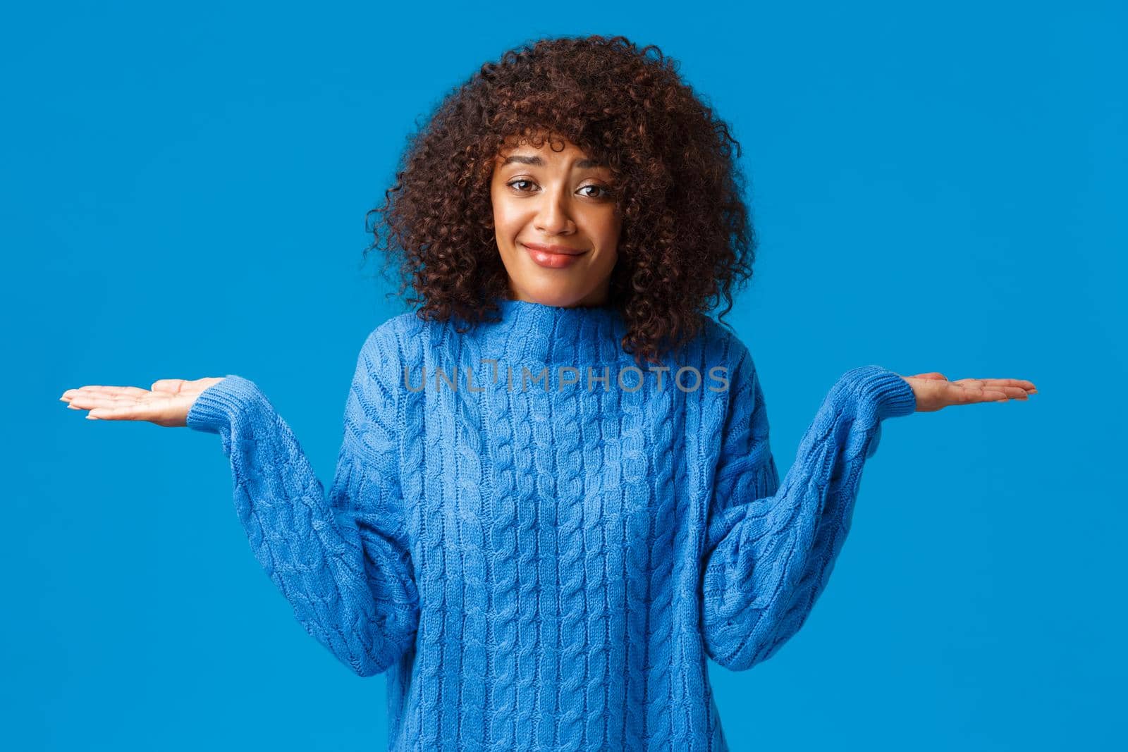 Who cares i dont know. Confused and clueless silly cute smiling african-american female in winter sweater, shrugging with hands spread sideways and smile as have no idea, standing blue background.