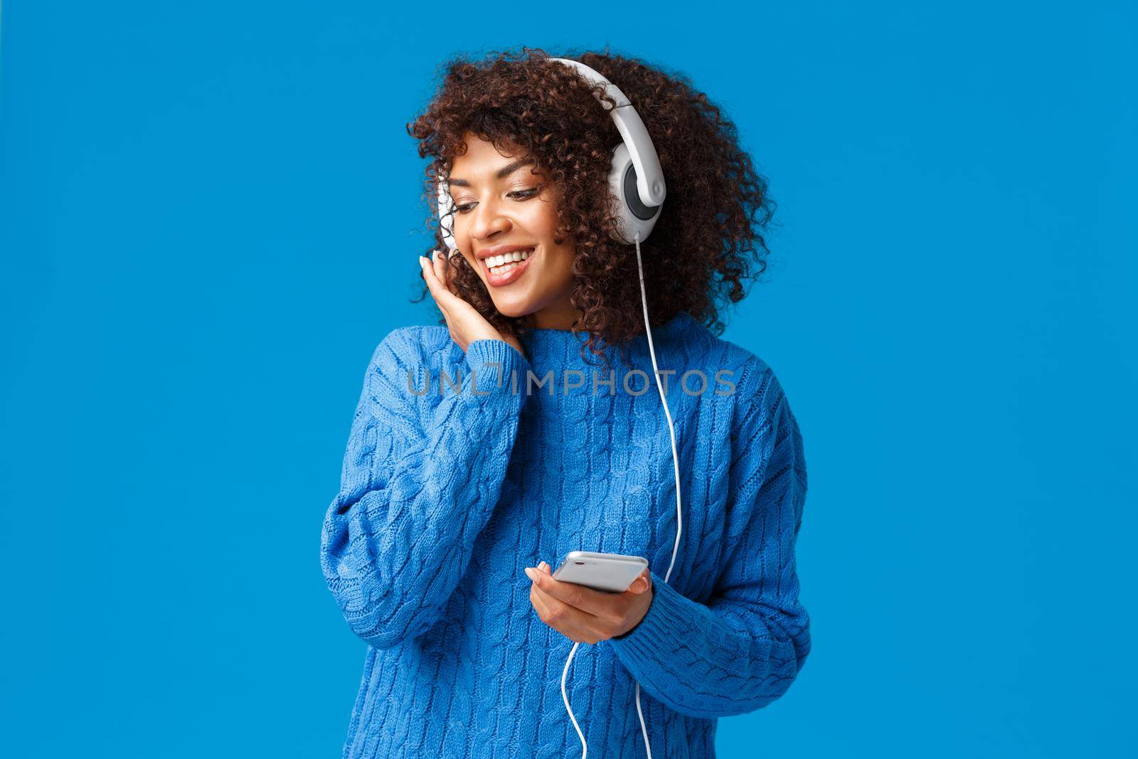 Lovely cute african-american female in winter sweater, listening romantic song, enjoying cool present headphones, touching earphones and gazing downstairs from window, smiling hold smartphone.