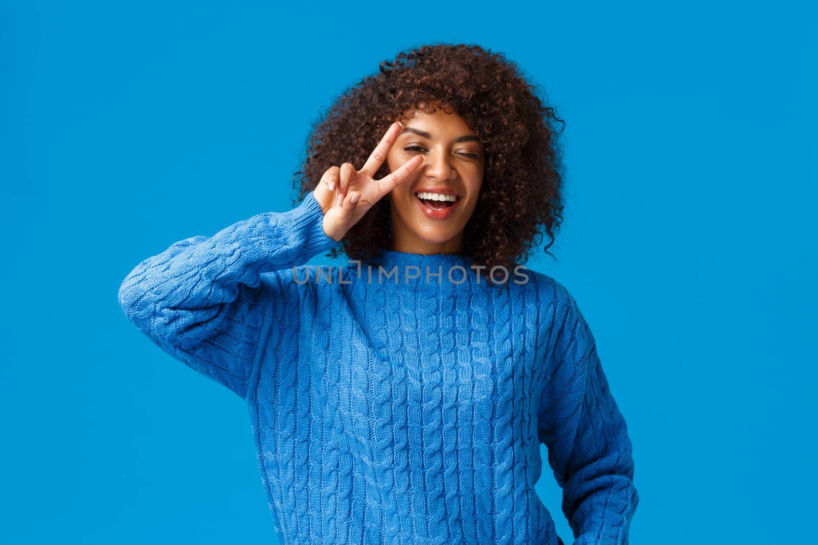 Carefree lovely, charismatic african american woman say cheeze posing delighted and upbeat, showing peace sign over winking eye and smiling, laughing happily, enjoy winter holidays, blue background.