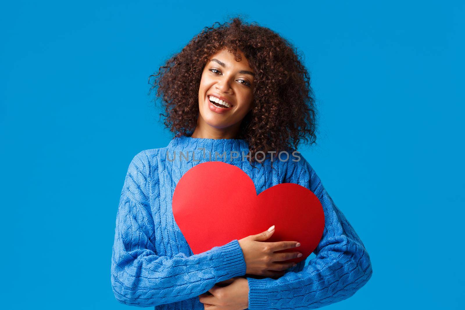 Cute and tender funny, smiling african-american female with afro haircut, press big red heart sign to chest and embrace it with delighted charming grin, showing love and affection, blue background by Benzoix