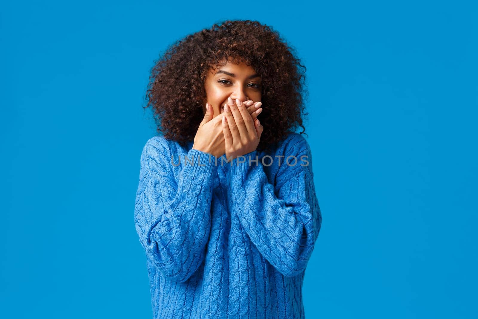 Cute and silly, lovely african-american woman with afro haircut, chuckling smiling and cover mouth as trying hold laugh, standing blue background making fun of friend, standing blue background by Benzoix