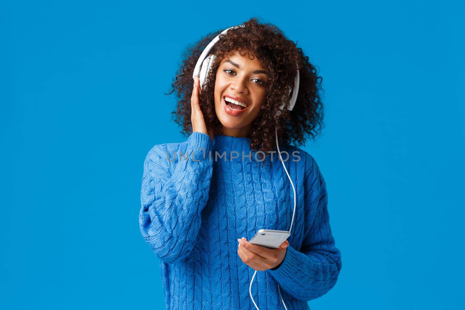 Happy and cheerful african-american woman playing karaoke game app in smartphone, wearing headphones singing and enjoying awesome music, sound quality, standing blue background.