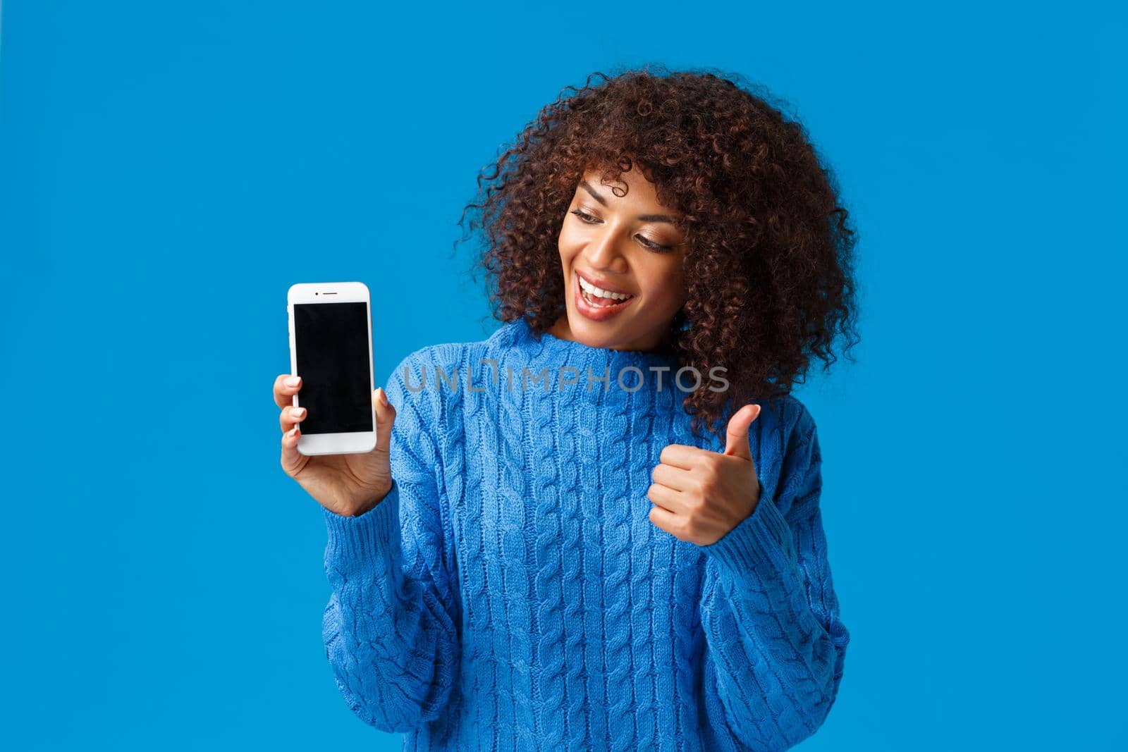 Cute and cheerful, satisfied african-american woman showing something on display, make thumb-up gesture, looking smartphone screen smiling in approval and delight, standing blue background.