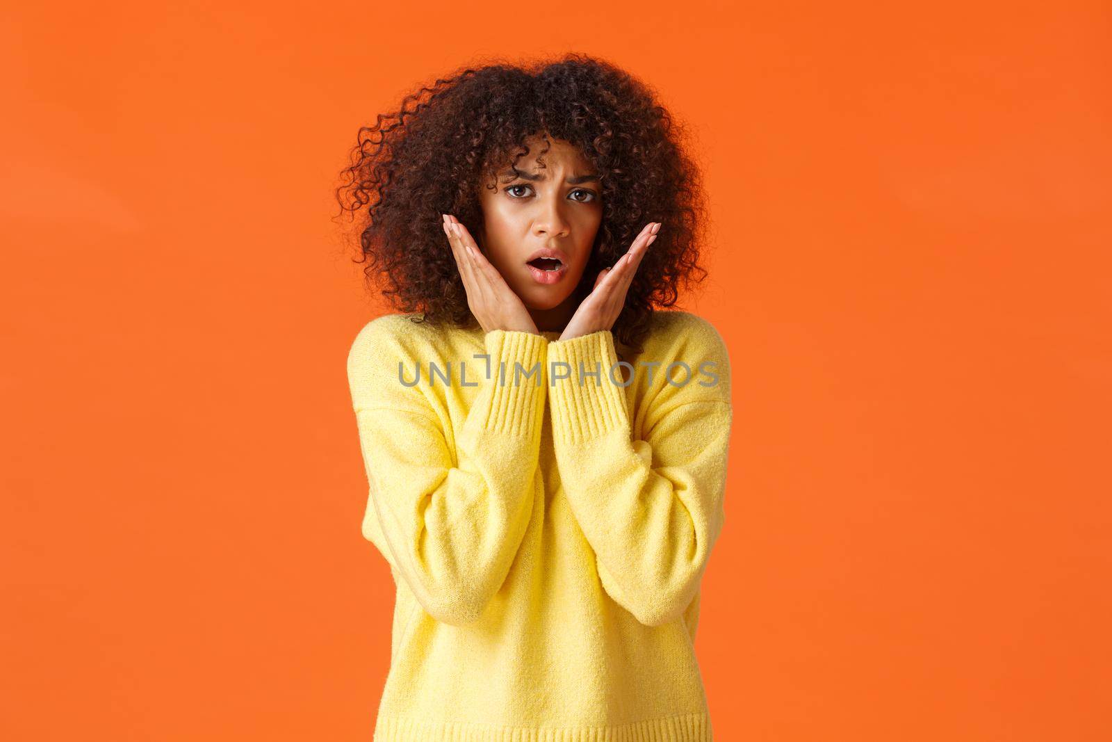 Scared timid and insecure lovely young girl with afro haircut, yellow sweater, gasping shocked and concerned, touching and staring in panic camera, feeling afraid, standing orange background.